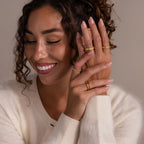 A smiling woman with curly hair shows off an Organic Fingerprint Band in 18K Gold on her finger while wearing a cream sweater.