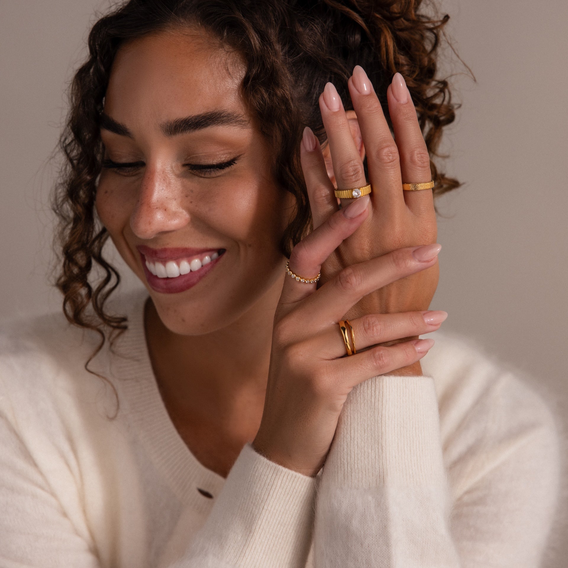 A smiling woman with curly hair shows off an Organic Fingerprint Band in 18K Gold on her finger while wearing a cream sweater.