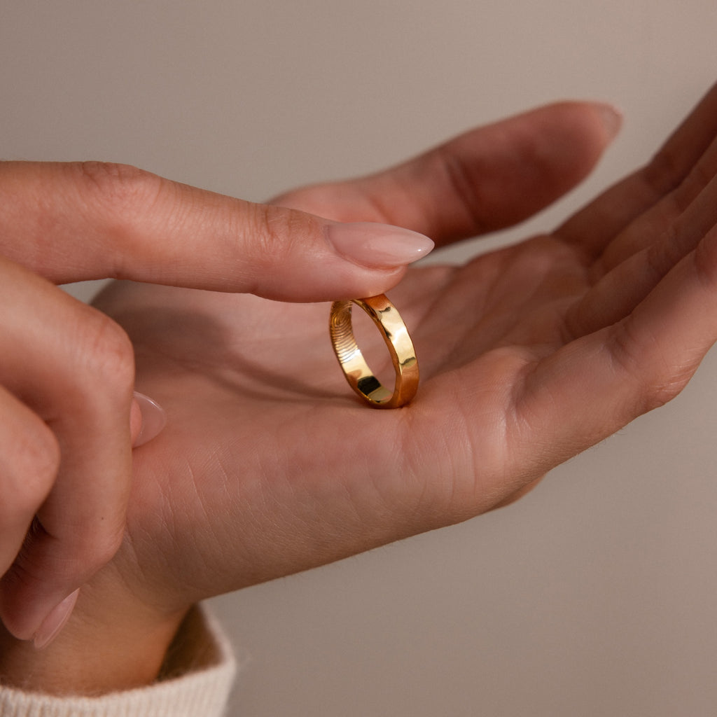 A hand holds the Organic Fingerprint Band in 18K Gold while another finger gently touches it, creating a tender moment against a neutral background.