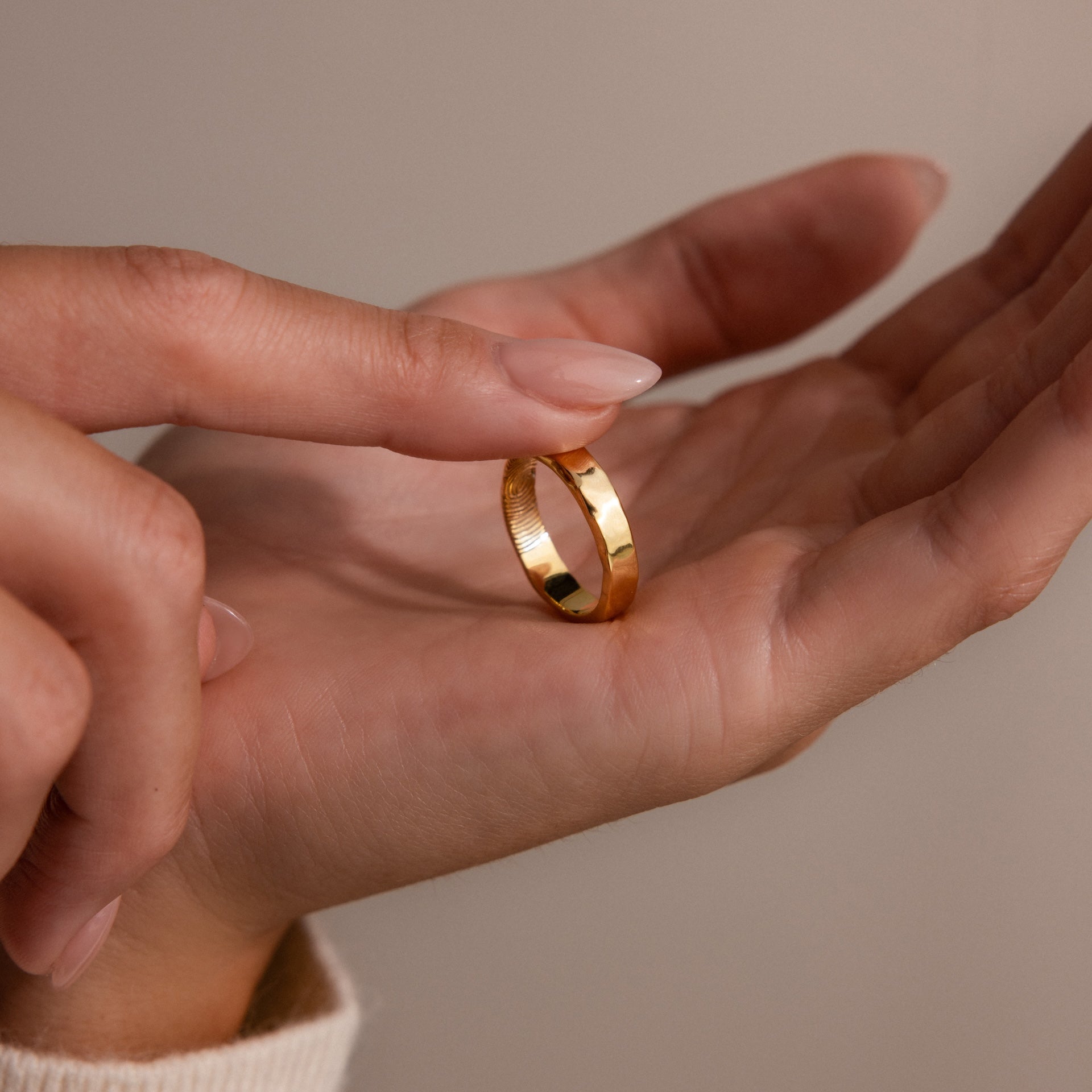 A hand holds the Organic Fingerprint Band in 18K Gold while another finger gently touches it, creating a tender moment against a neutral background.