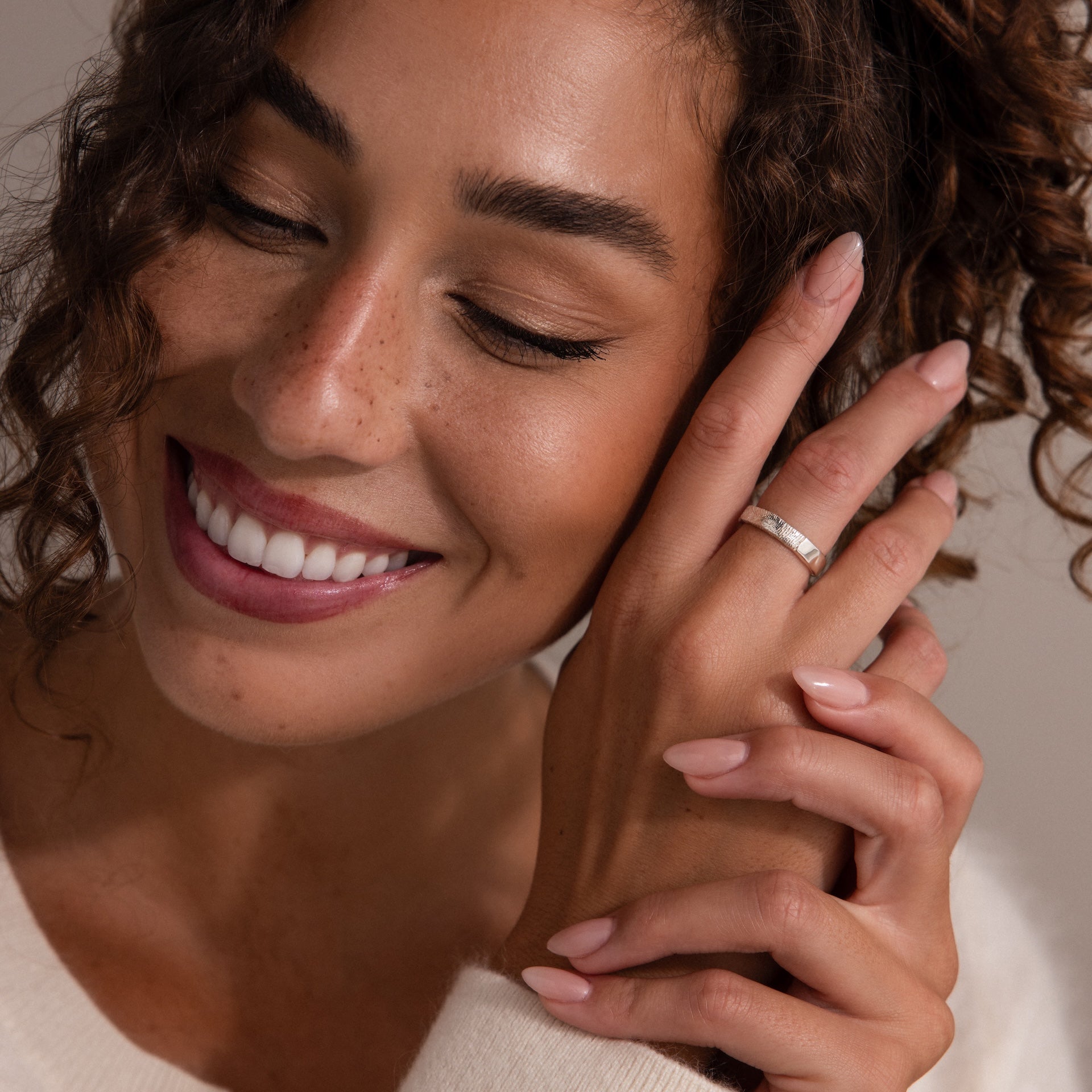 Smiling woman with curly hair shows off an Organic Fingerprint Band on her finger, wearing a cream sweater.