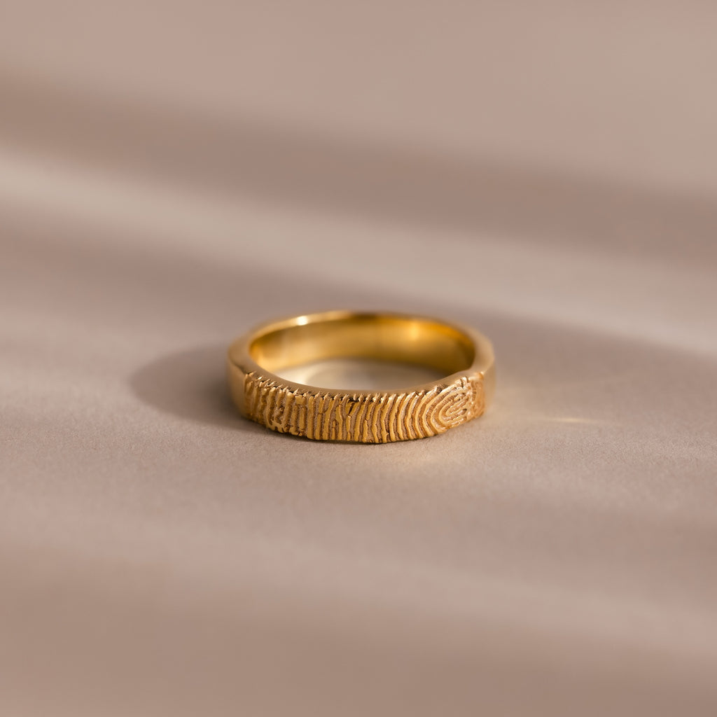 A custom-engraved Organic Fingerprint Band in 18K Gold rests on a smooth, beige surface—perfect as a memorial keepsake.