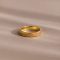 A custom-engraved Organic Fingerprint Band in 18K Gold rests on a smooth, beige surface—perfect as a memorial keepsake.