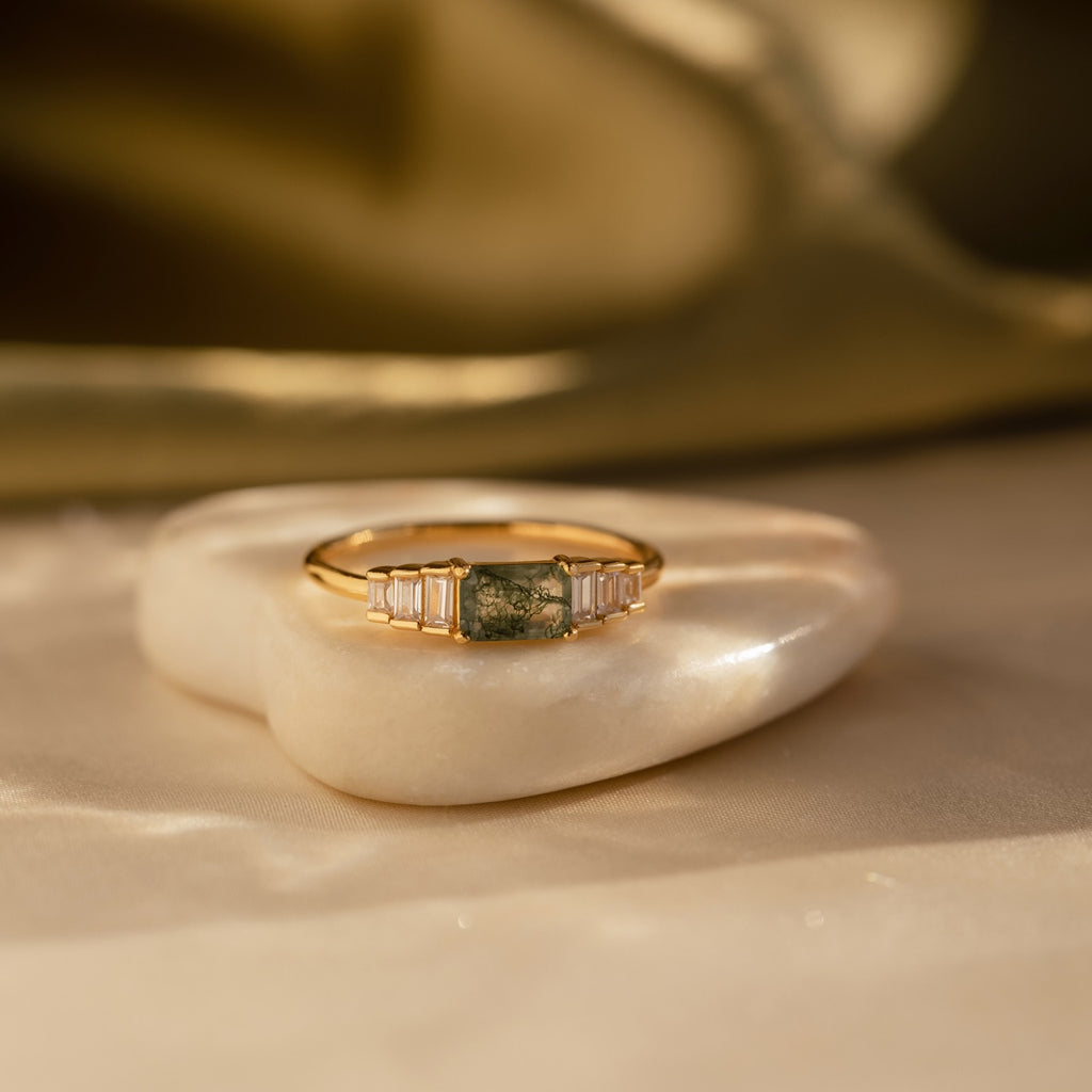 Gold moss agate ring resting on a smooth, heart-shaped white stone, bathed in warm light that reflects off the gemstone and gold setting.
