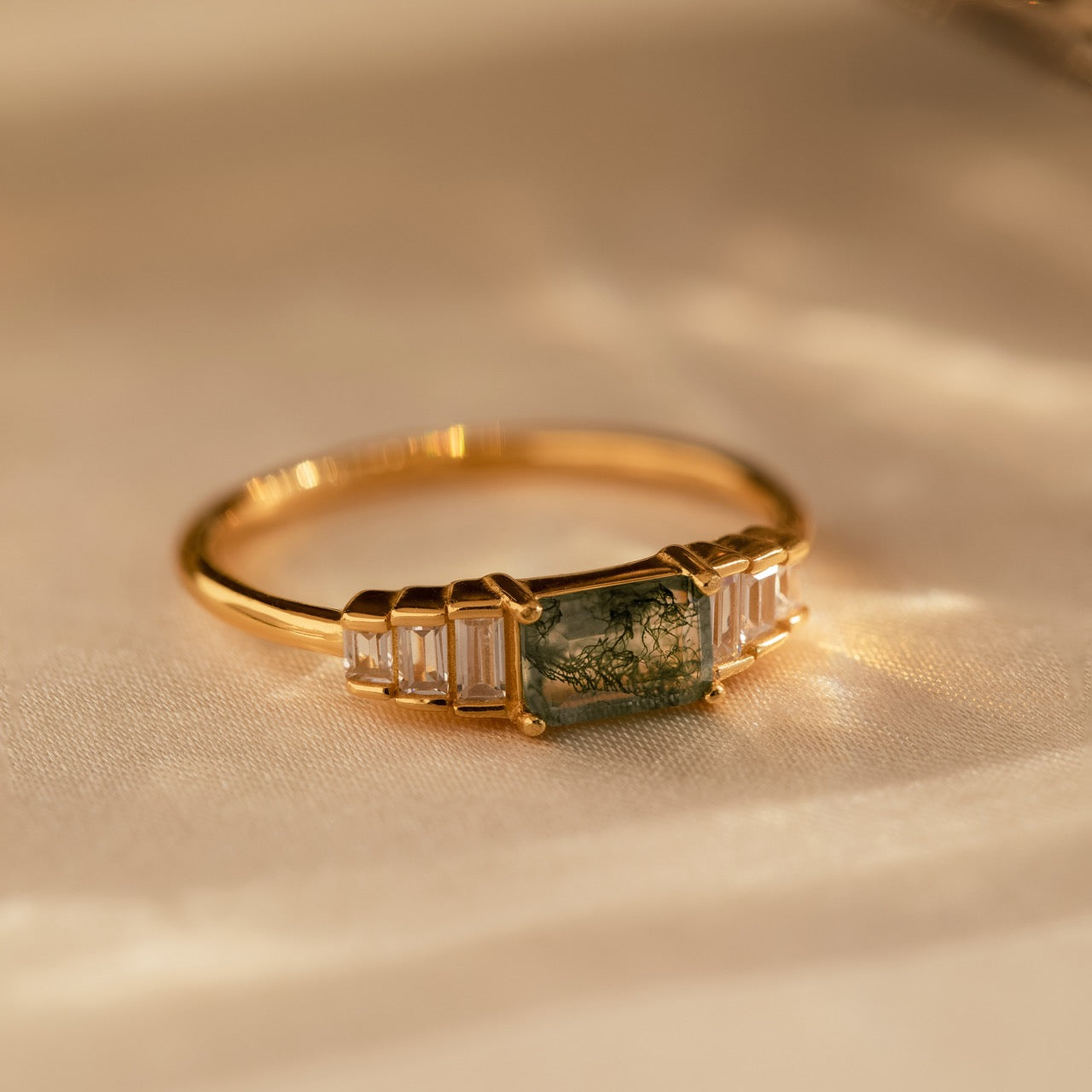 Gold moss agate ring displayed flat on soft champagne-toned fabric, highlighting the rich green inclusions of the center stone and the clear baguette-cut accents.
