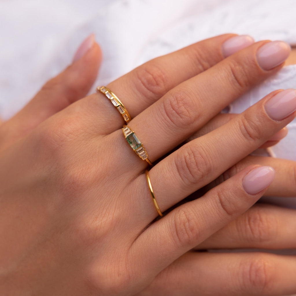 Hand wearing the moss agate ring alongside a slim plain gold band and a matching baguette-style band, styled with light pink manicured nails.