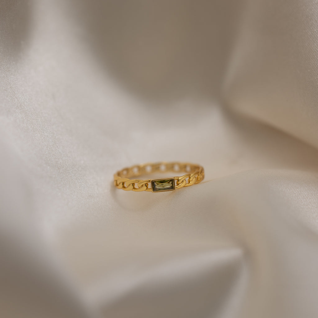 The Baguette Peridot Curb Ring, highlighted by its bezel-set gemstone, is displayed on soft, creamy draped fabric.