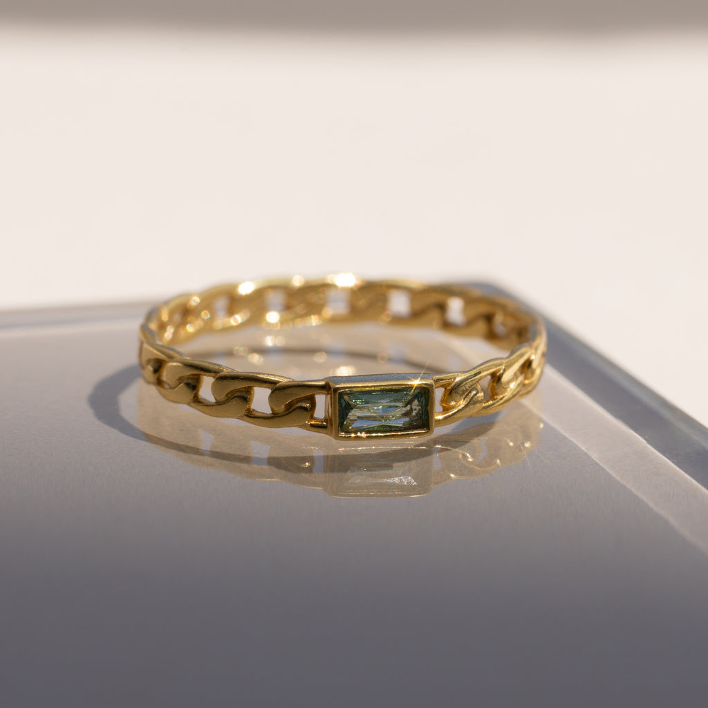 The Baguette Peridot Curb Ring features a rectangular bezel-set gemstone on a reflective surface in soft light. Its elegant curb-link band lends a modern edge to this striking piece.