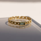 The Baguette Peridot Curb Ring features a rectangular bezel-set gemstone on a reflective surface in soft light. Its elegant curb-link band lends a modern edge to this striking piece.