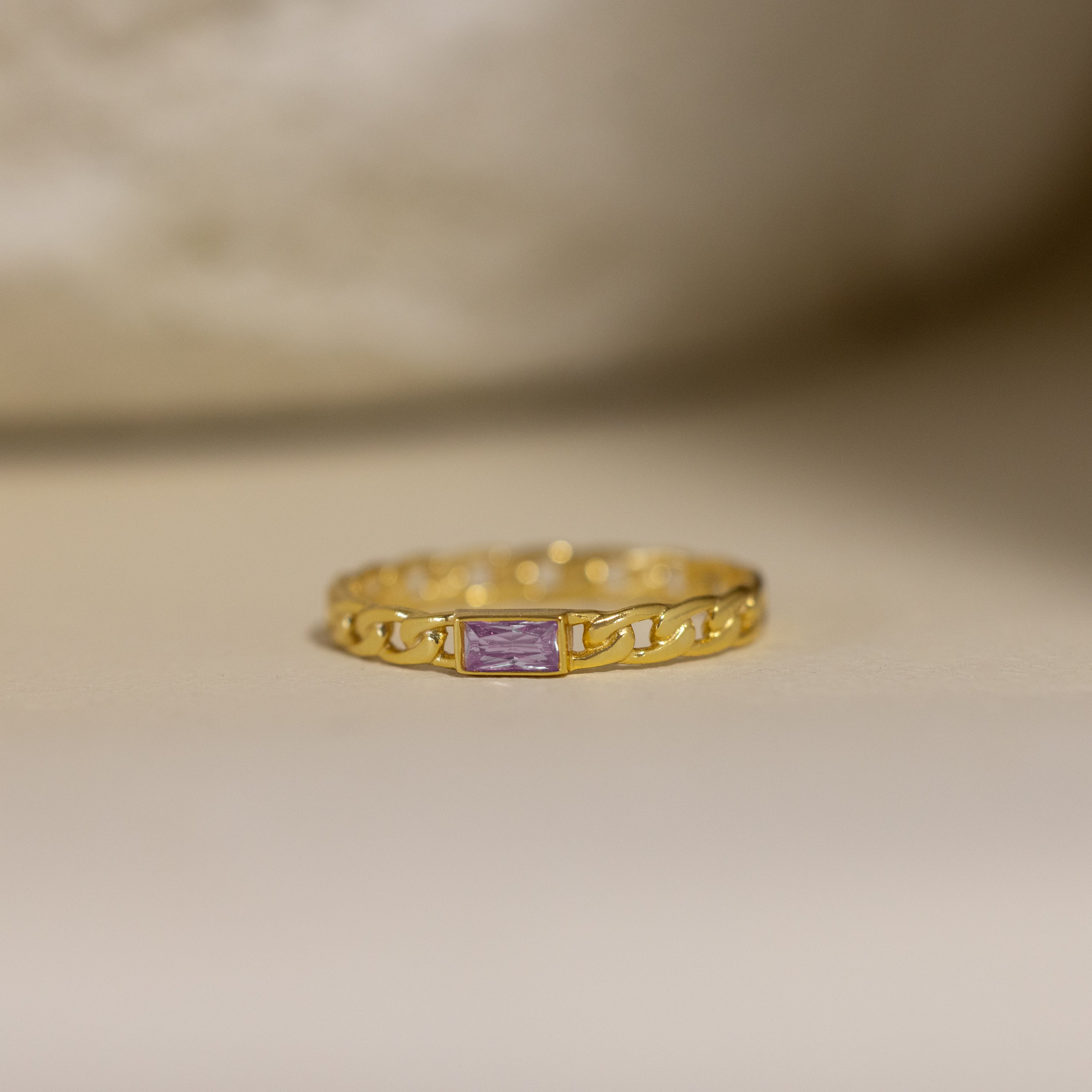 The Baguette Alexandrite Curb Ring features a gold band with a rectangular purple stone and a delicate curb-link design, displayed on a neutral surface.