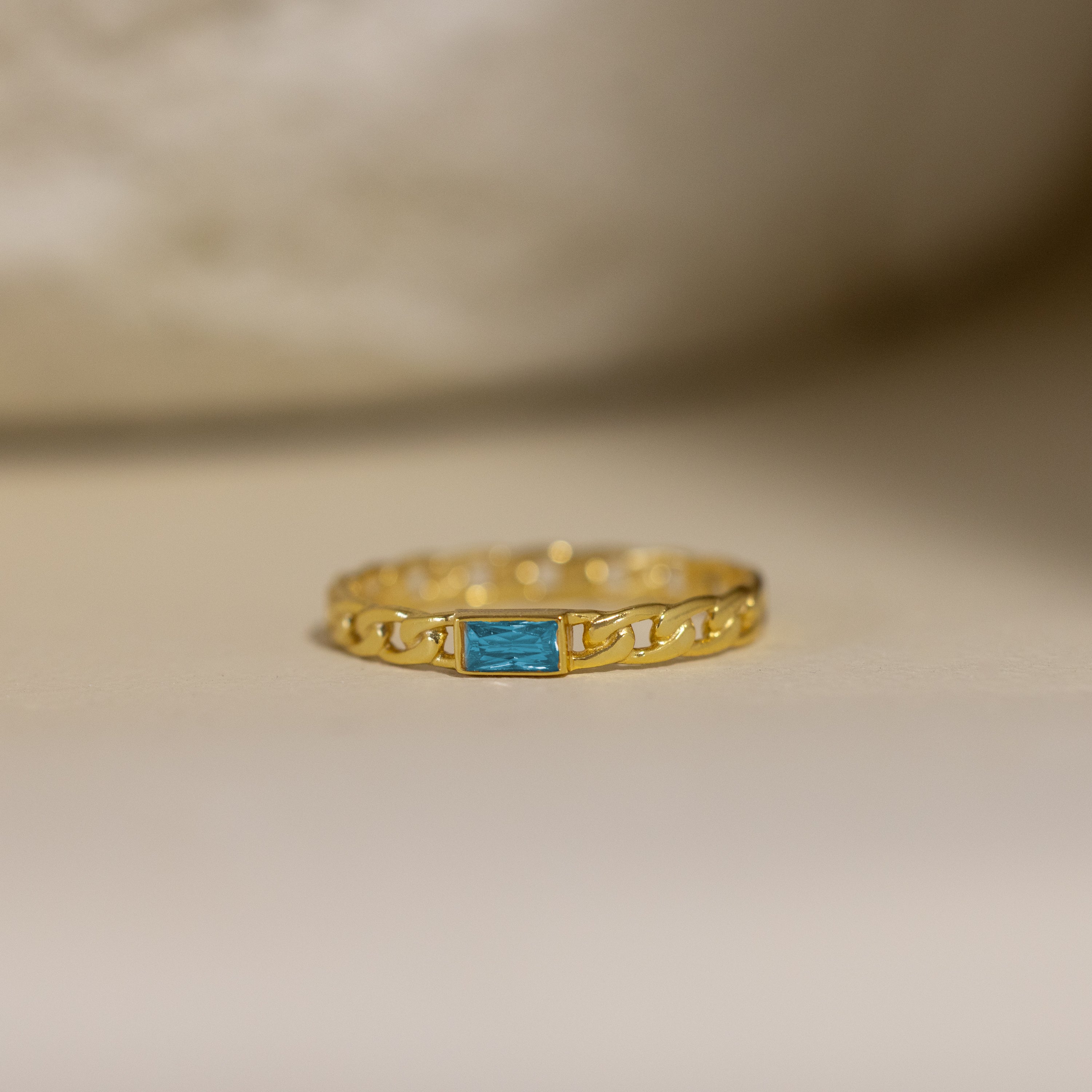 The Baguette Blue Zircon Curb Ring features a striking rectangular gemstone and an elegant curb-link band, displayed on a neutral background—ideal for anyone who enjoys personalized jewelry.
