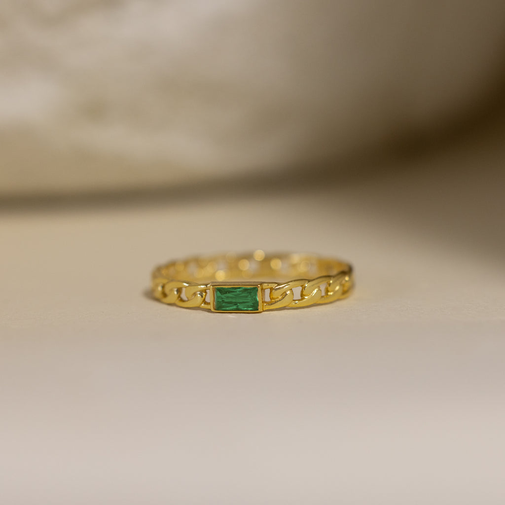 The Baguette Emerald Curb Ring features a rectangular bezel-set emerald on a curb-link band, showcased against a neutral background.