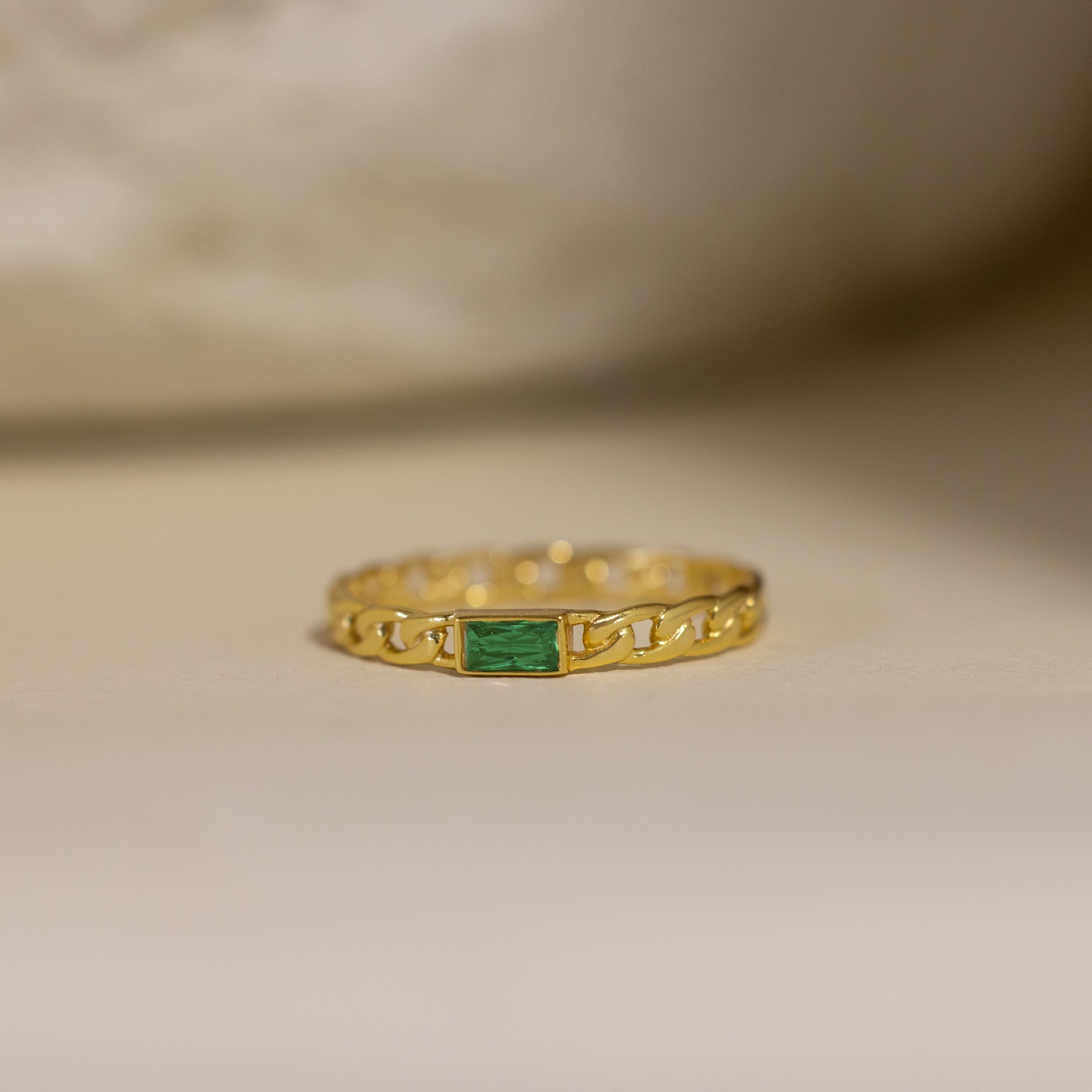 The Baguette Emerald Curb Ring features a rectangular bezel-set emerald on a curb-link band, showcased against a neutral background.