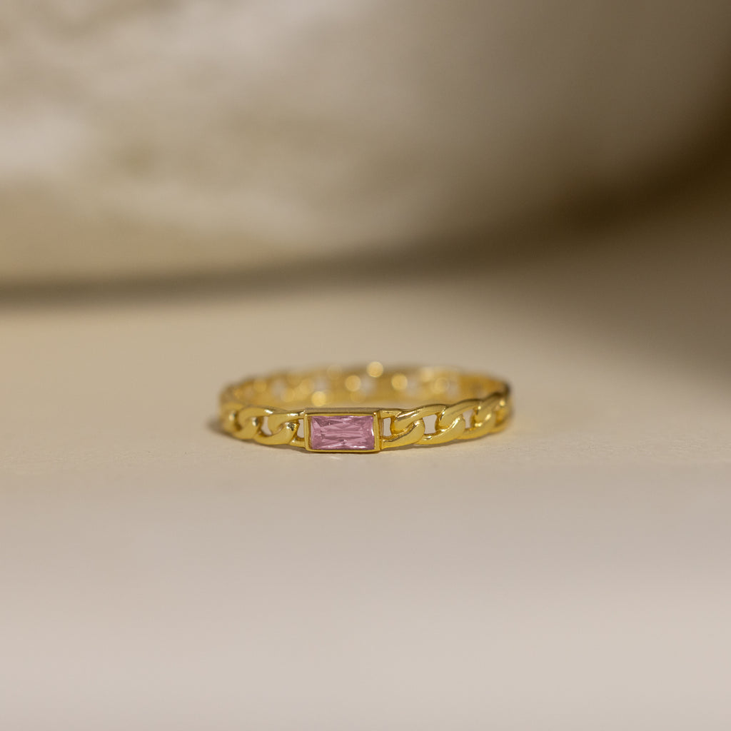 The Baguette Pink Tourmaline Curb Ring features a pink tourmaline baguette on a delicate, chain-inspired gold band, set against a soft background.