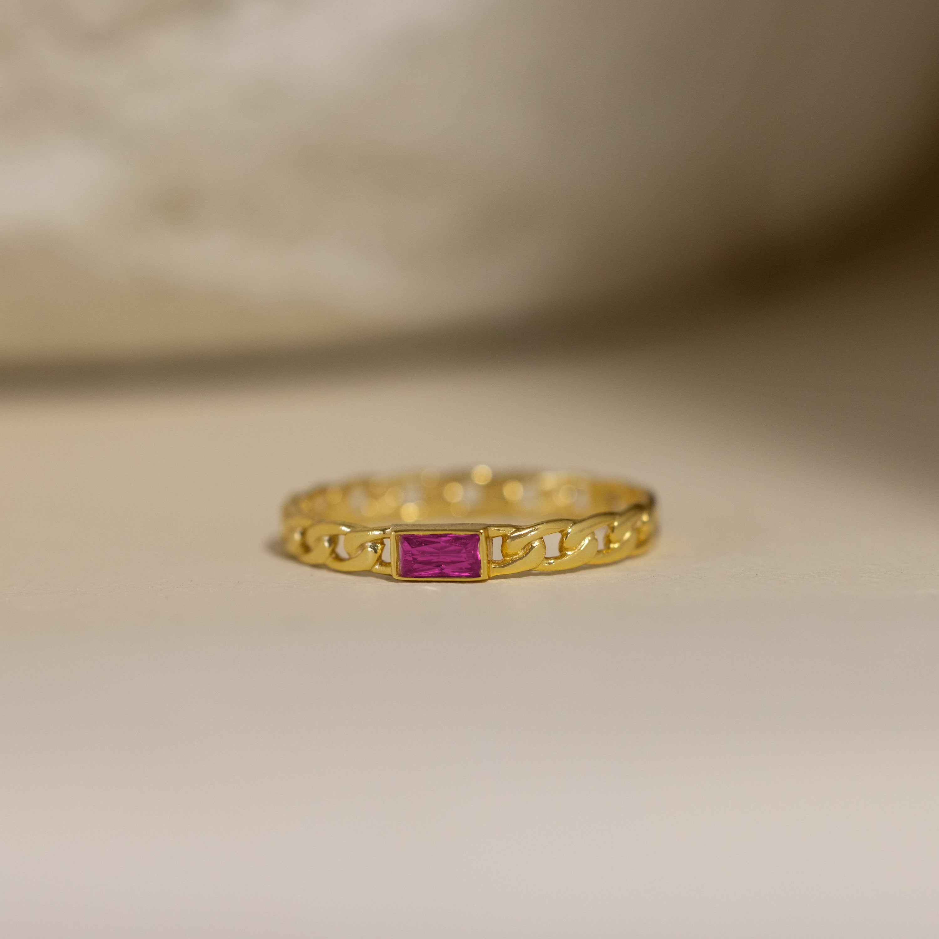 The Baguette Ruby Curb Ring features a gold curb-link band set with a rectangular pink gemstone, displayed on a neutral background.