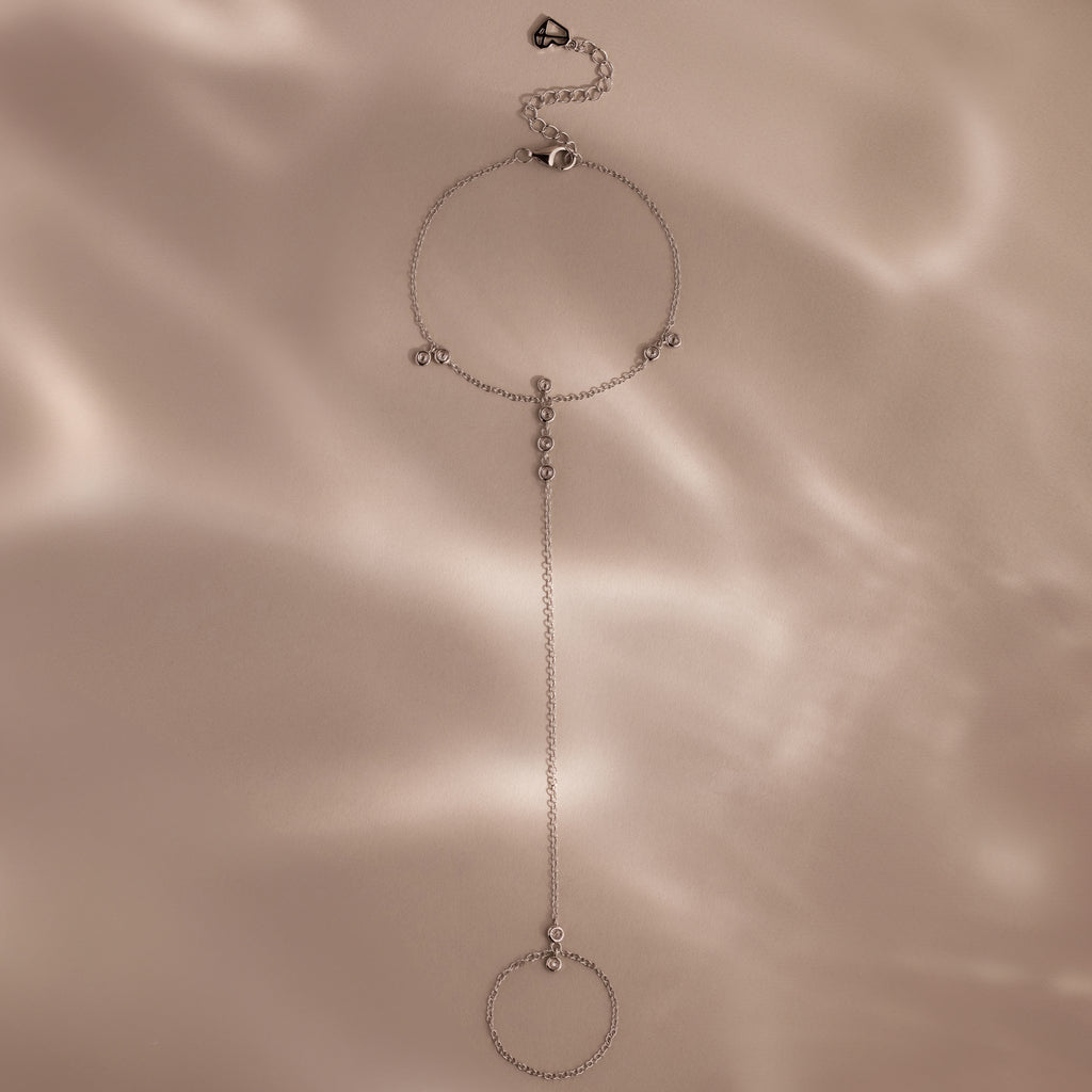 Delicate silver hand chain bracelet with small bezel-set stones displayed on a soft beige background with gentle shadows.