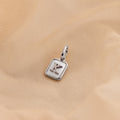 A silver square pendant charm with a raised letter 'K' on a soft beige fabric background.