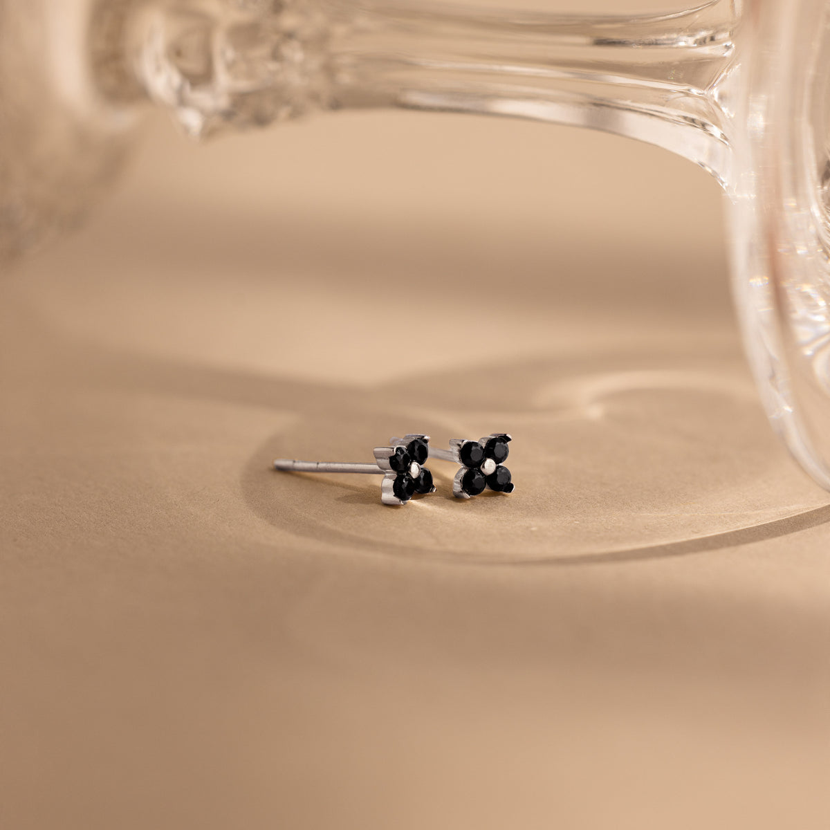 A pair of small black flower stud earrings with silver posts, resting on a beige surface with a soft-focus glass object in the background.