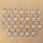 Twenty silver letter charms spelling out the alphabet, arranged in four rows of five on a textured beige background.