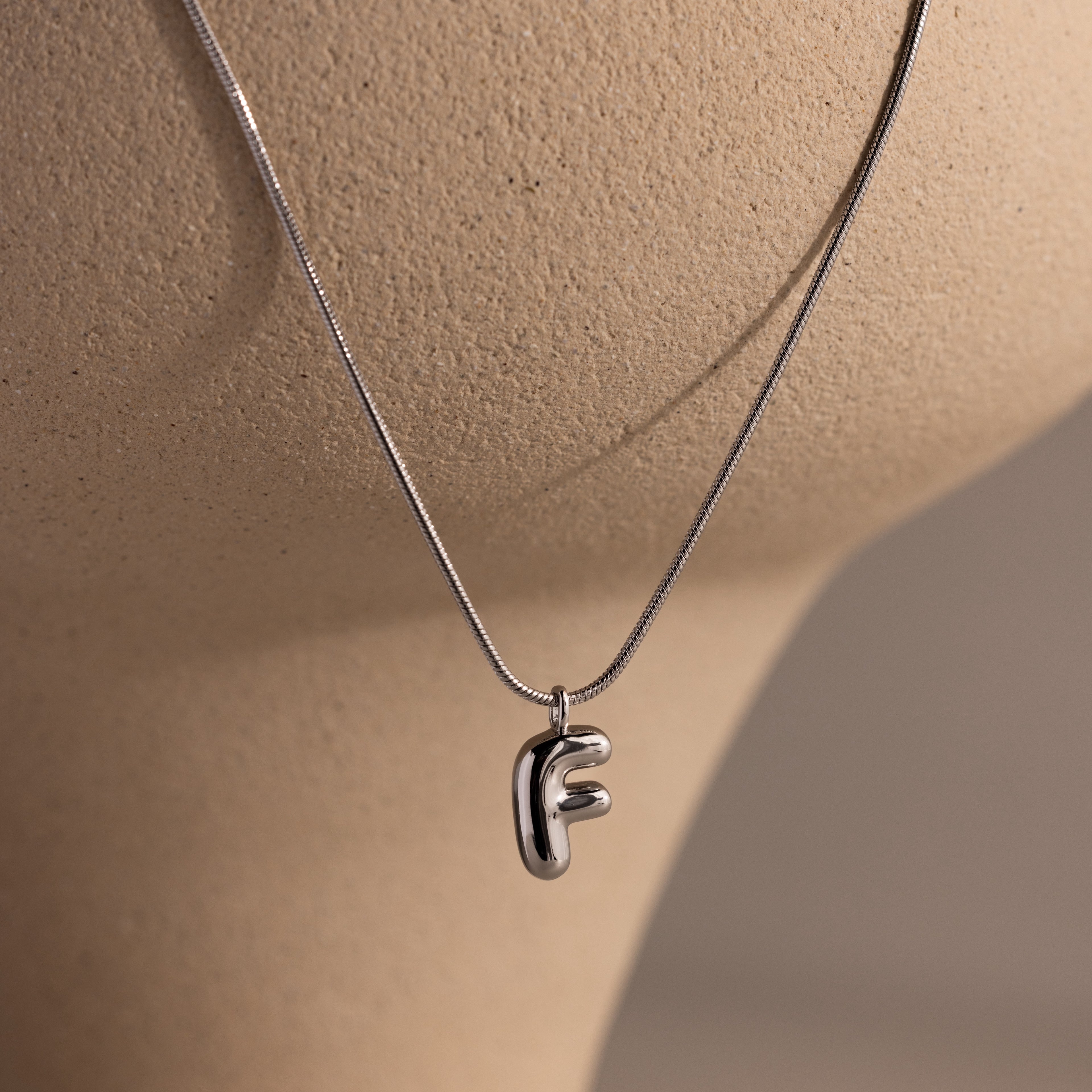 A Bubble Letter Necklace in White Gold featuring a metallic letter F pendant is displayed on a beige mannequin, making it ideal for fans of personalized jewelry.