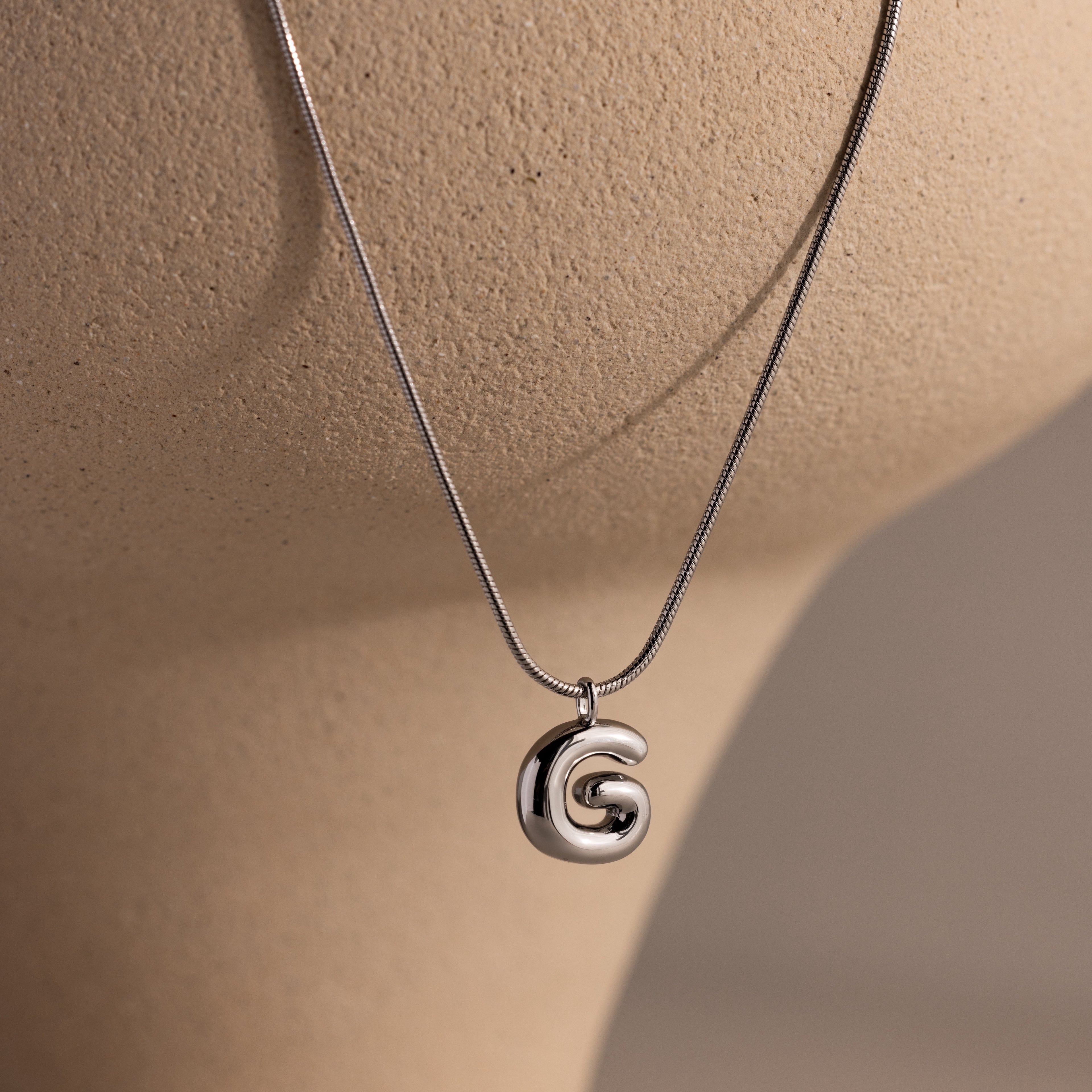 The Bubble Letter Necklace in White Gold features a chunky, shiny G pendant displayed on a beige mannequin, making it the perfect personalized accessory for any outfit.