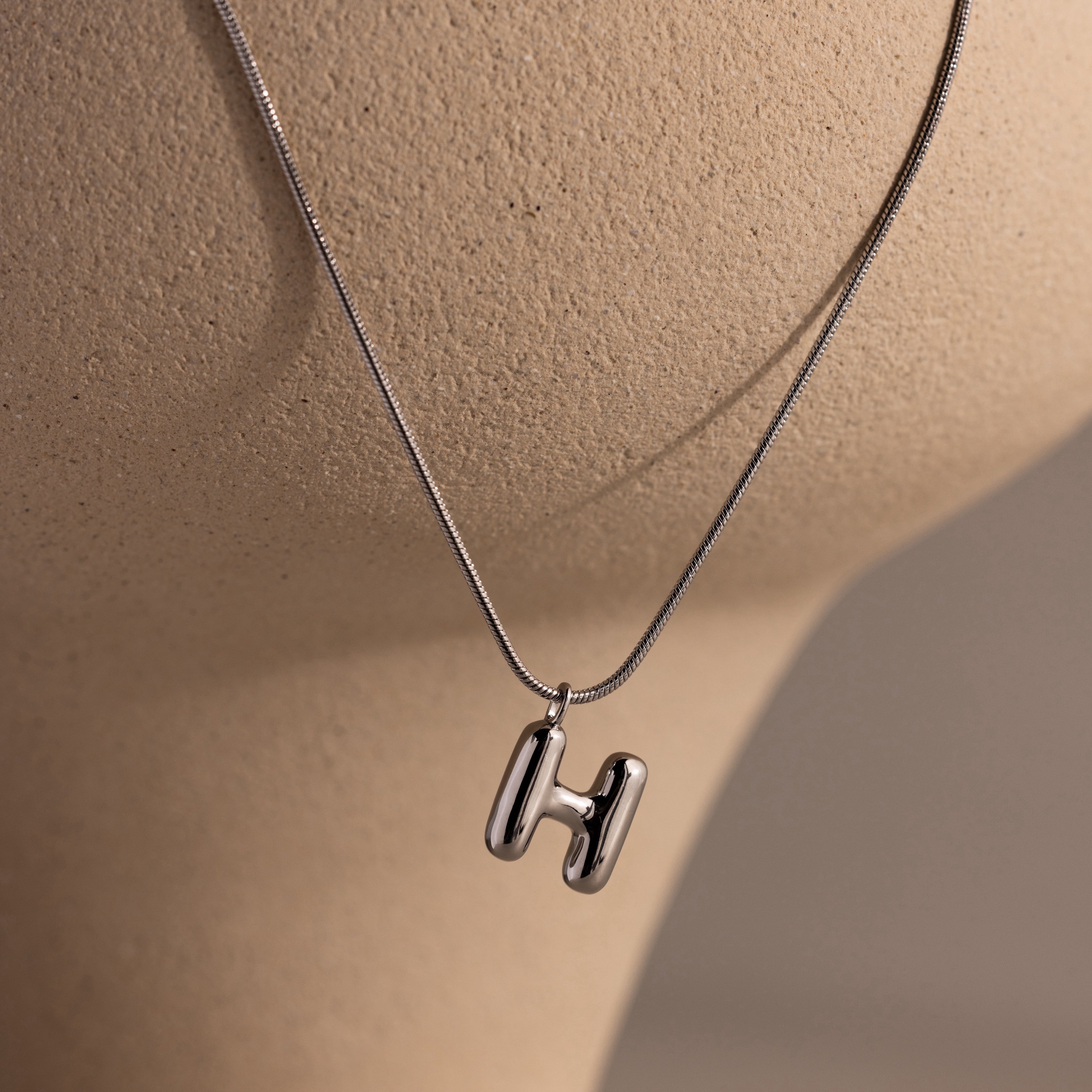A Bubble Letter Necklace in white gold featuring a chunky H pendant is showcased on a beige mannequin—an ideal choice for fans of personalized jewelry.