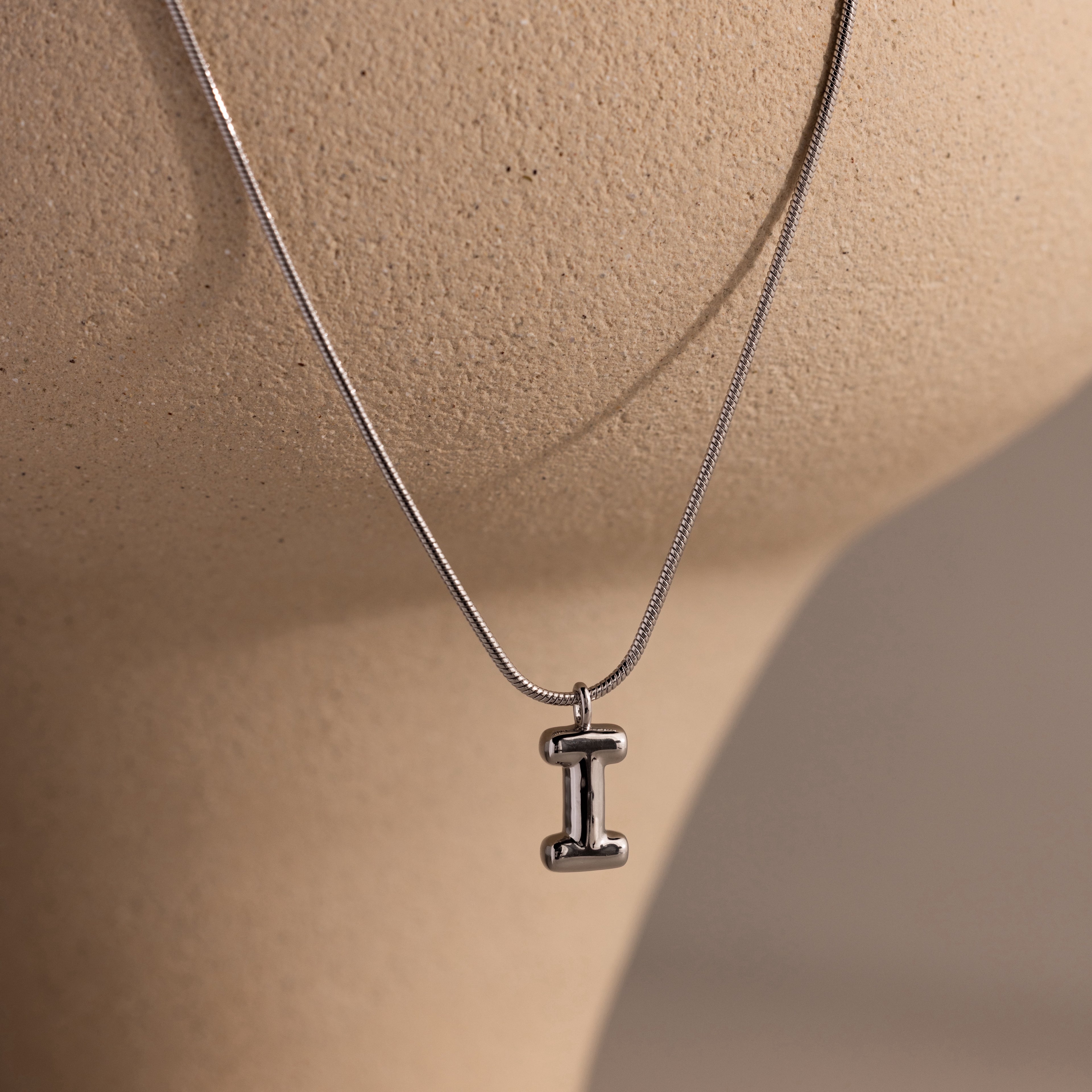 The Bubble Letter Necklace in White Gold features a chunky pendant shaped like the letter I, displayed on a beige textured surface—ideal for anyone who loves personalized jewelry.