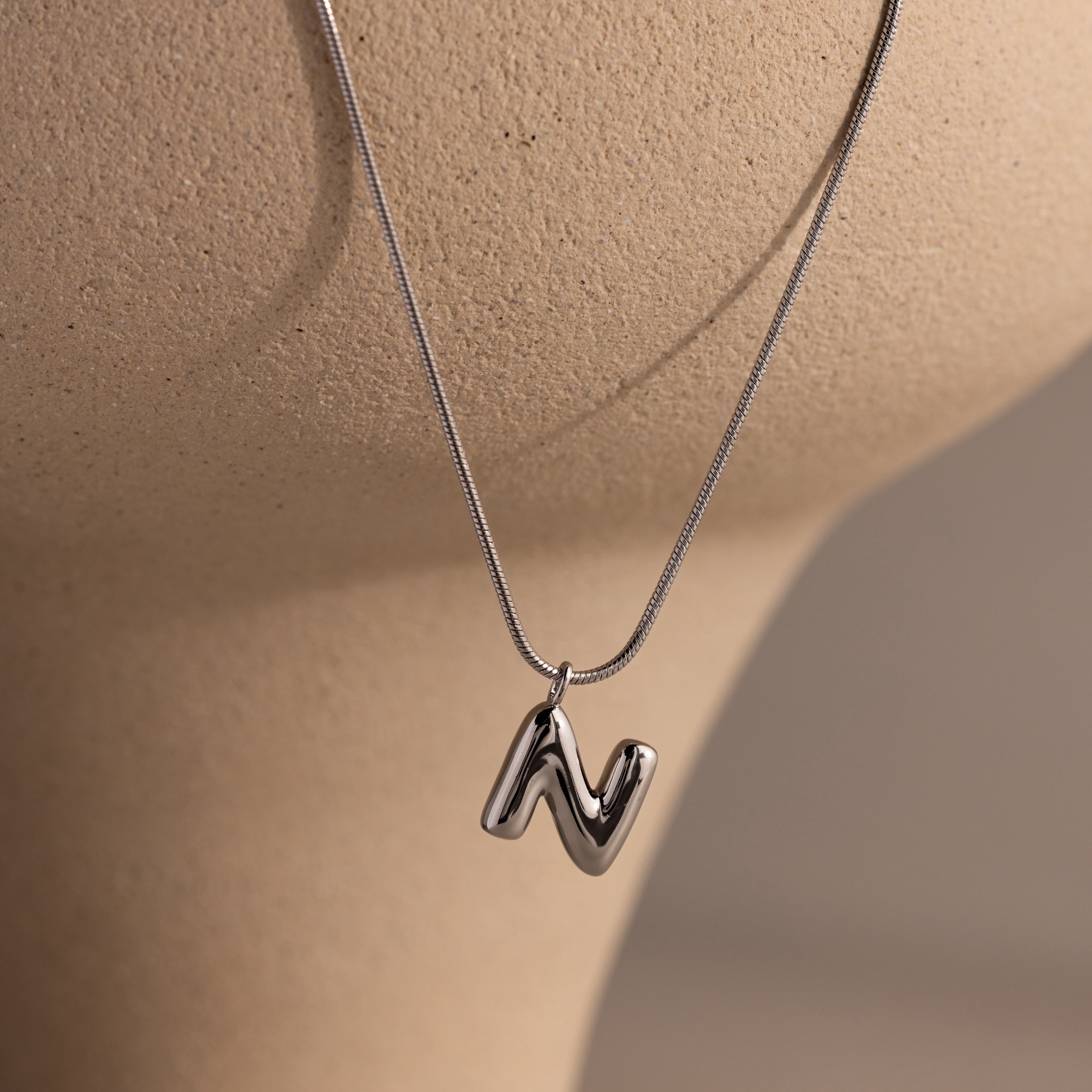 A Bubble Letter Necklace in White Gold with a shiny N pendant rests against a beige textured surface—perfect for lovers of personalized jewelry.