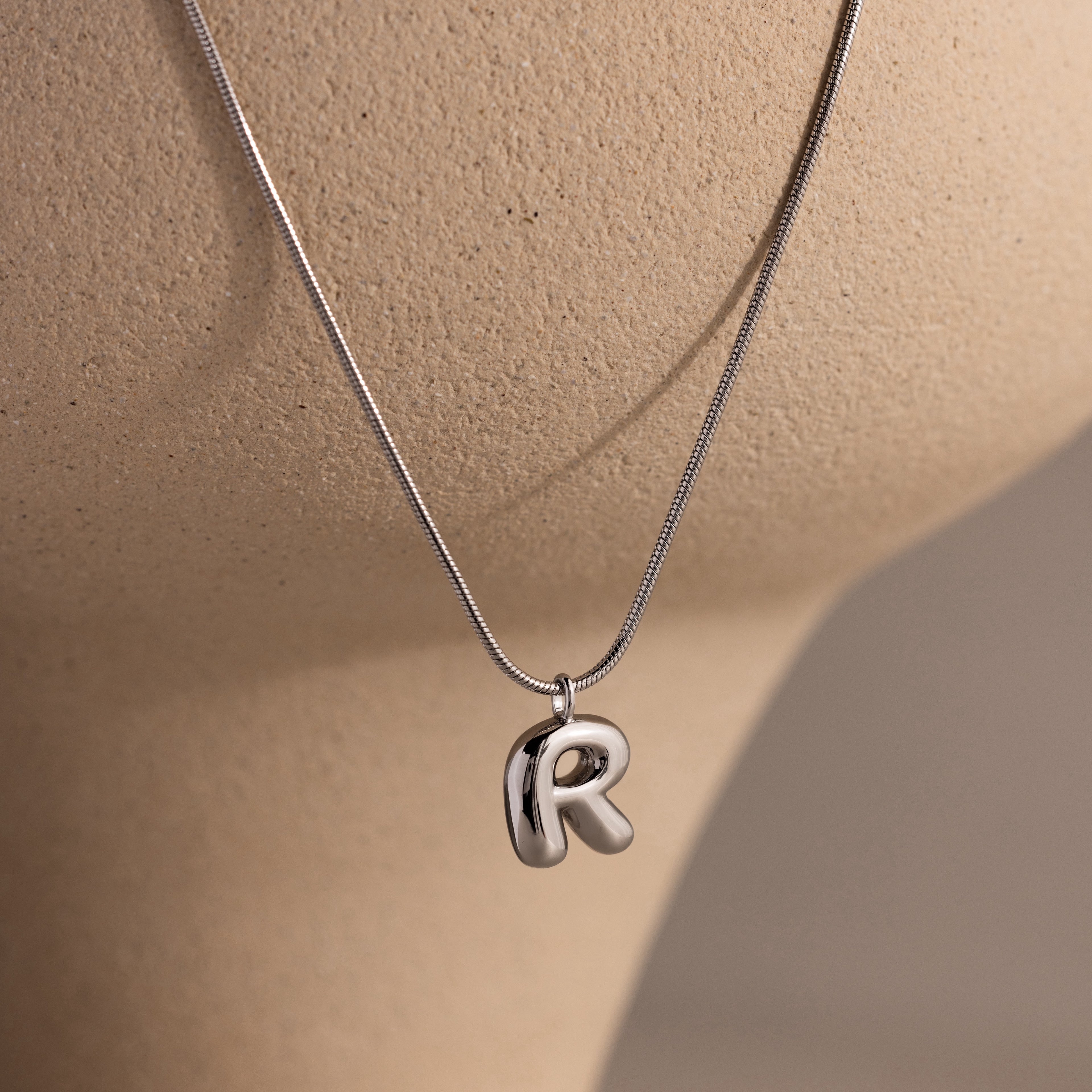 The Bubble Letter Necklace in White Gold features a shiny letter R pendant, displayed on a beige mannequin—ideal for fans of personalized jewelry.