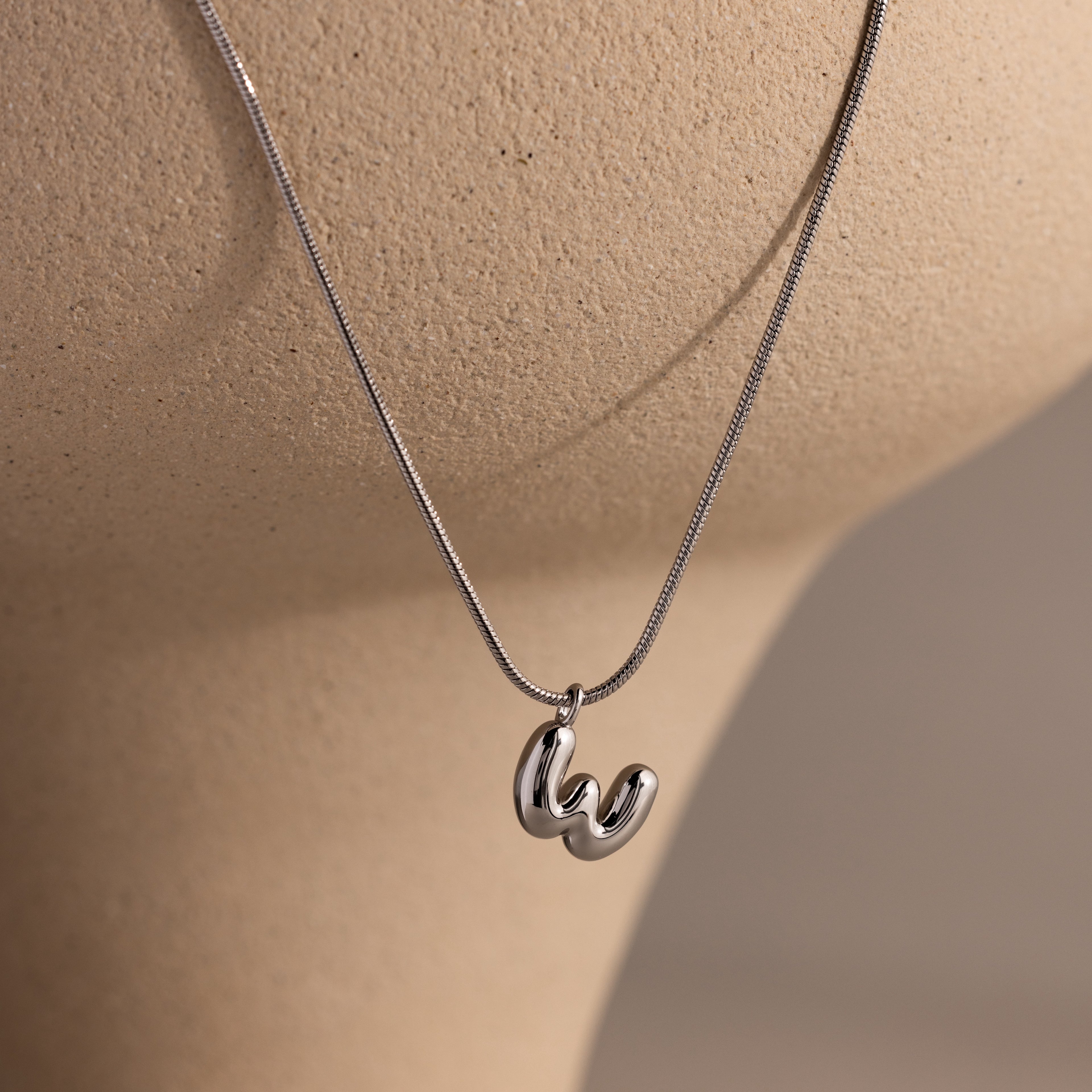 The Bubble Letter Necklace in White Gold features a chunky silver lowercase "w" pendant on a beige mannequin bust—ideal for fans of personalized jewelry.