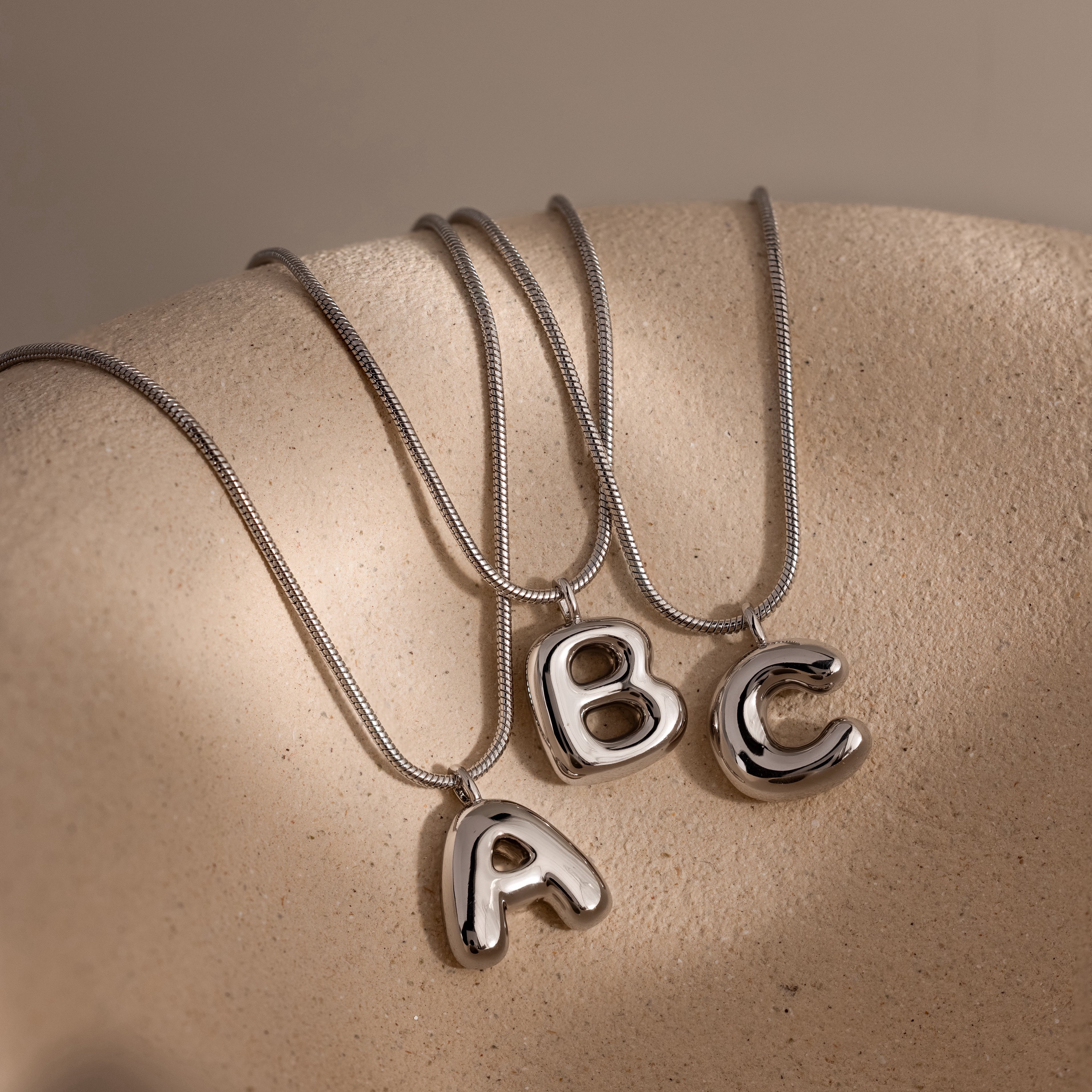 The Bubble Letter Necklace in White Gold features bold A, B, or C pendants with a chunky design, displayed on a textured beige surface—ideal for those seeking modern, personalized jewelry.