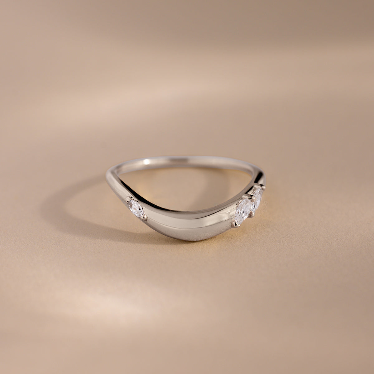 A silver teardrop-shaped ring with three clear gemstones, photographed on a warm beige background.