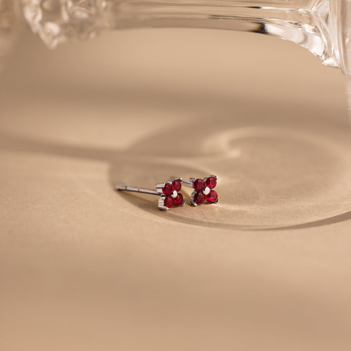 A pair of small silver stud earrings with four pink ruby gemstones arranged in a flower-like cluster, photographed on a warm beige surface with a clear glass object partially visible at the top.