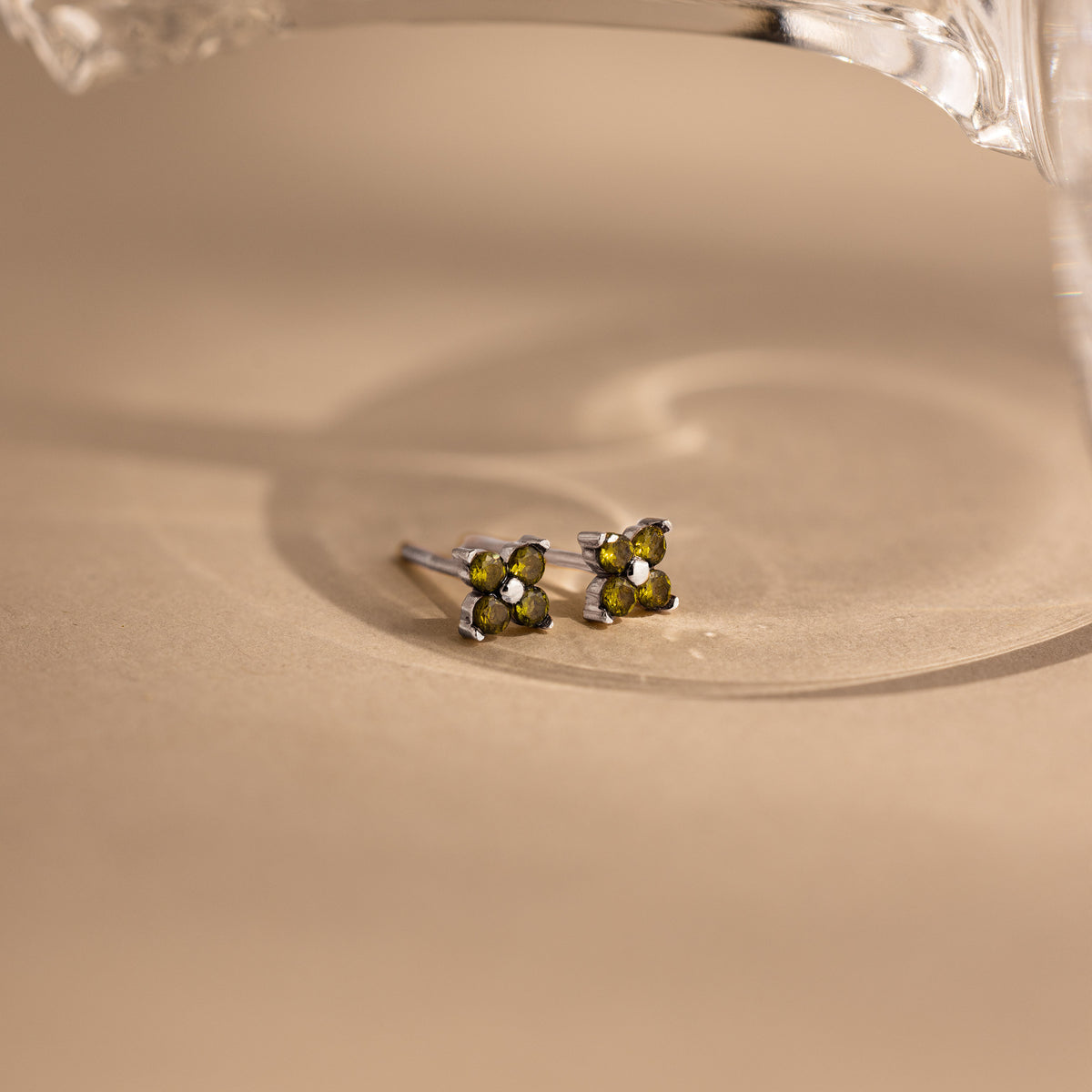 A pair of small silver stud earrings with four olive green gemstones arranged in a flower shape, resting on a beige surface with a soft shadow.