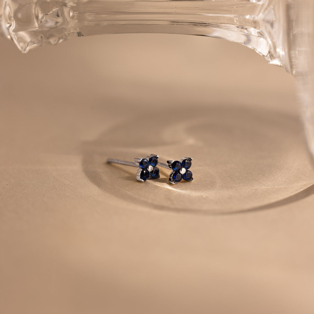 A pair of silver stud earrings with small flower-shaped blue gemstone clusters, resting on a beige surface under a clear glass object.