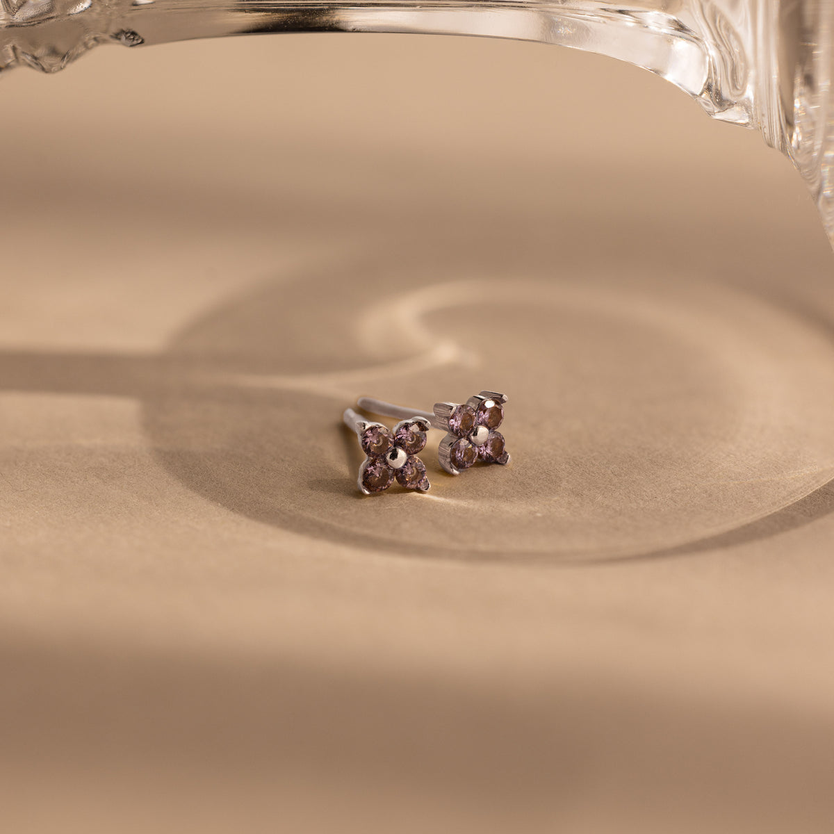 A pair of silver-toned stud earrings with four small light purple gemstones arranged in a flower shape, resting on a warm beige surface with soft shadows and a glass object partially visible at the top.
