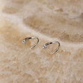 A pair of small silver open hoop earrings, each set with a single clear gemstone, displayed on a textured beige stone surface.