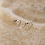 A pair of small silver open hoop earrings, each set with a single clear gemstone, displayed on a textured beige stone surface.