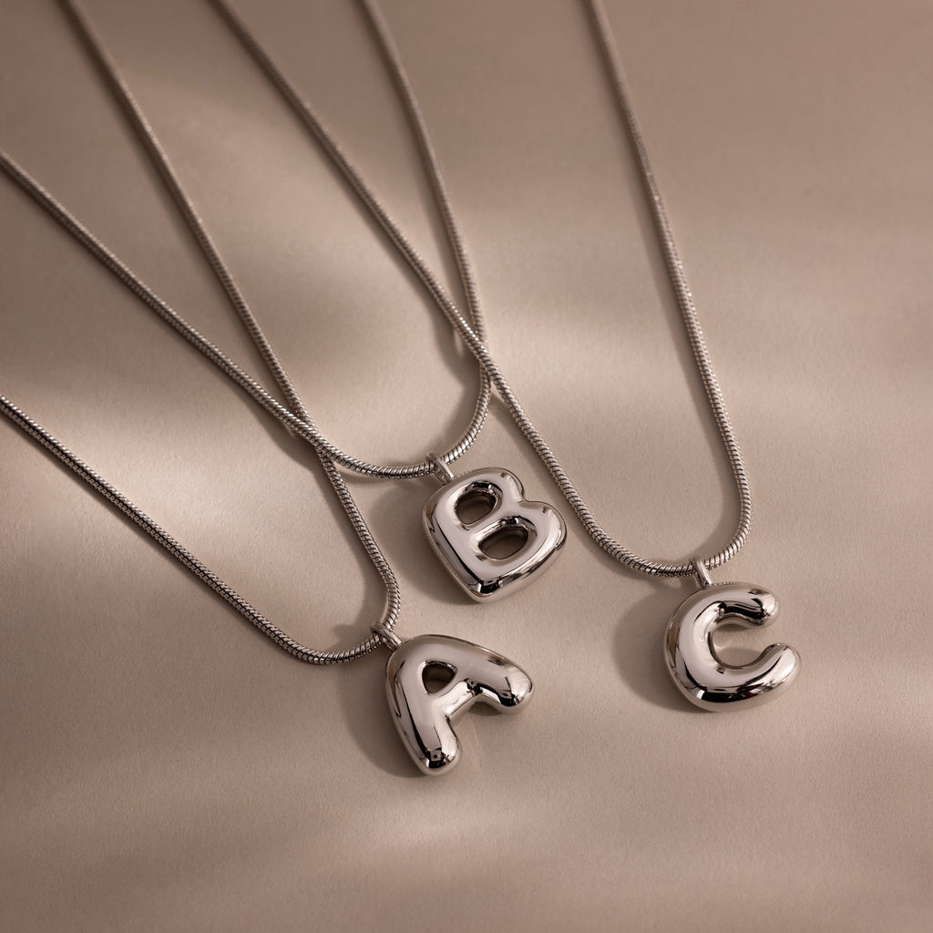 Three Bubble Letter Necklaces in White Gold, featuring shiny pendant letters A, B, and C, rest on a smooth beige surface—a chic choice for personalized jewelry lovers.