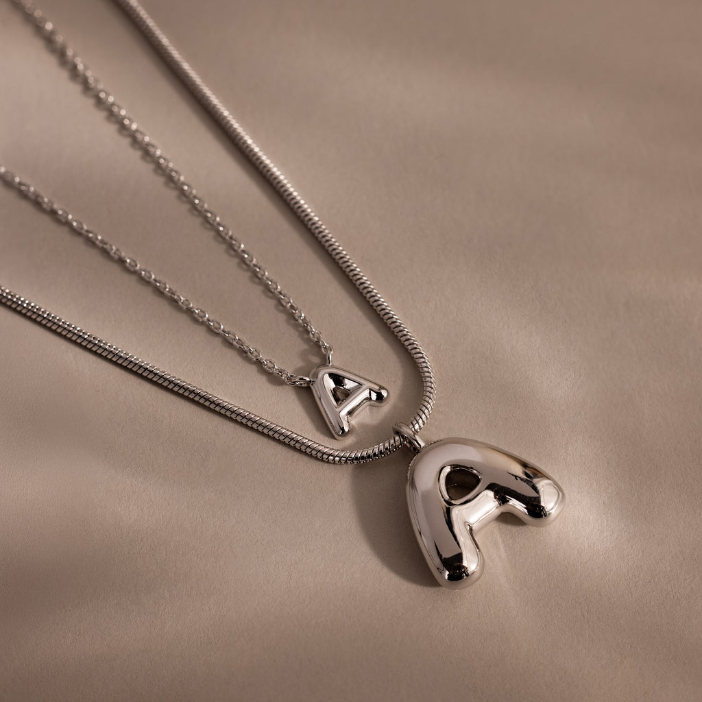 Two Dainty Bubble Letter Necklaces in Sterling Silver with shiny letter A pendants rest on a soft beige background—an elegant, personalized accessory for any style.