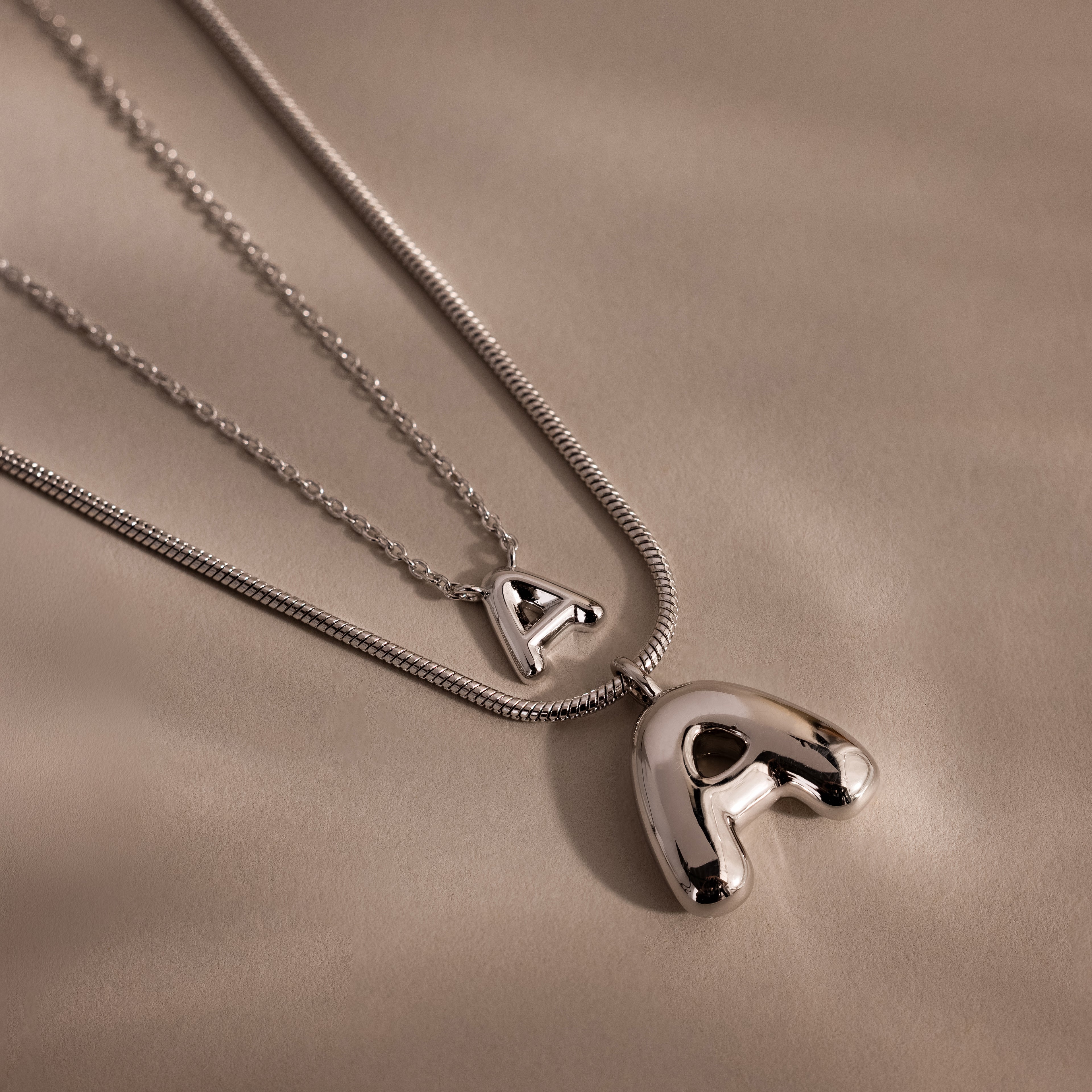 Two Dainty Bubble Letter Necklaces in Sterling Silver with shiny letter A pendants rest on a soft beige background—an elegant, personalized accessory for any style.