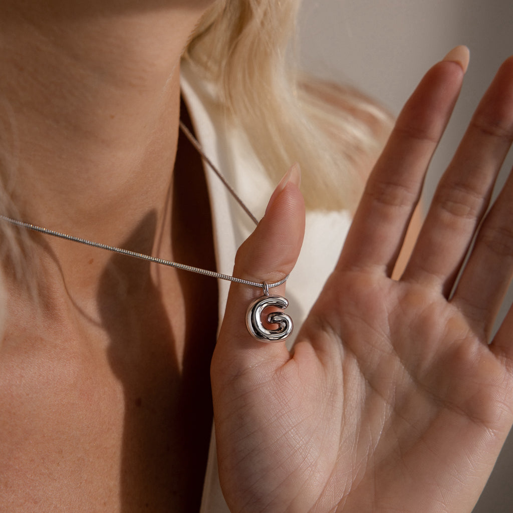 A hand holds the Bubble Letter Necklace near a woman's neck, highlighting its trendy silver interlocking loop design and personalized style.
