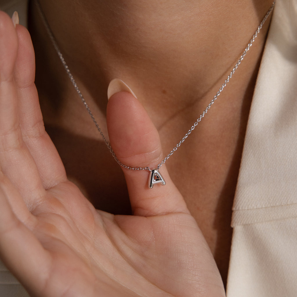 A hand holds the Dainty Bubble Letter Necklace in Sterling Silver, featuring a minimalist silver letter pendant, against a person's neck.