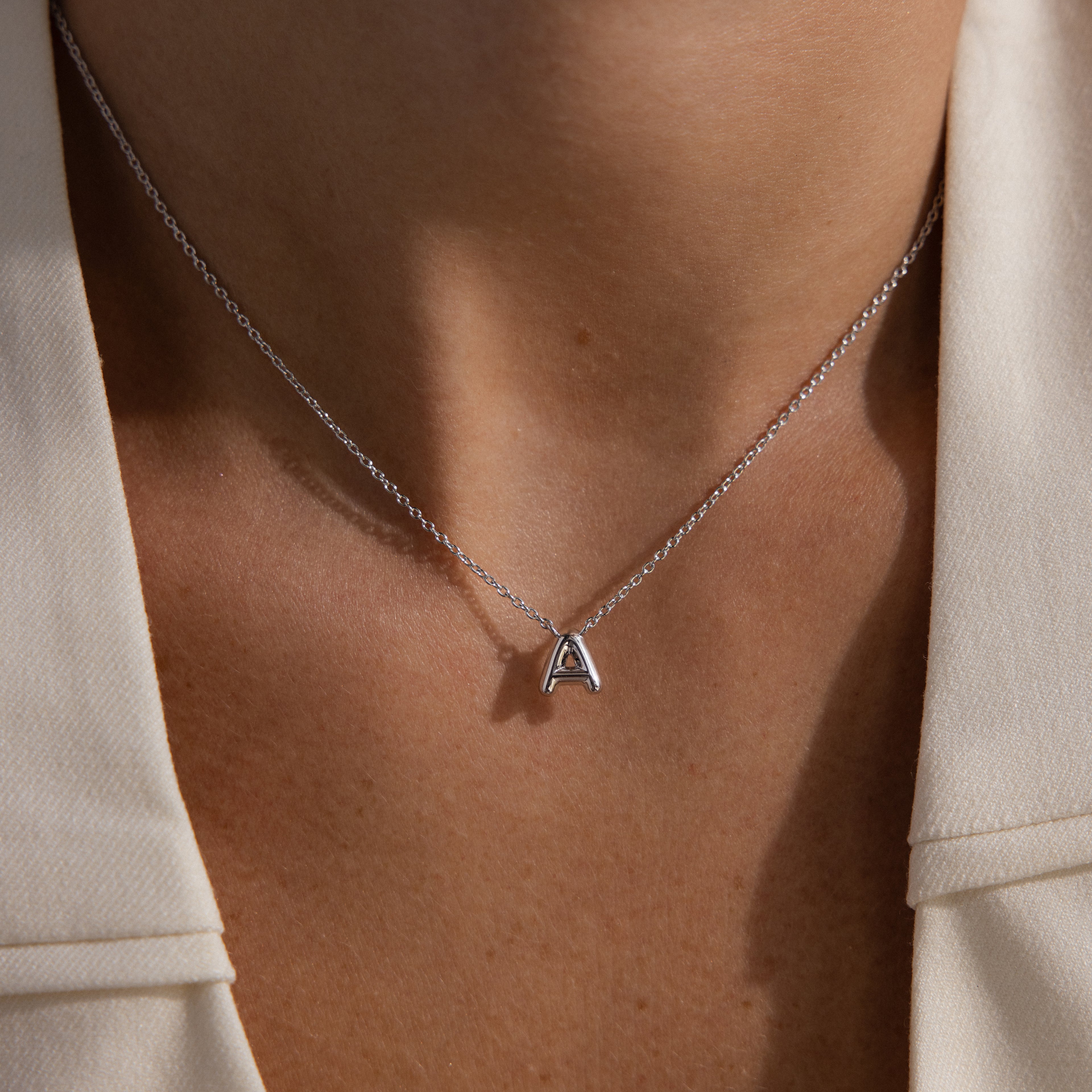 Wearing the Dainty Bubble Letter Necklace in Sterling Silver with an "A" pendant, beautifully paired with a cream-colored collared top.