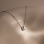 The Dainty Bubble Letter Necklace in Sterling Silver features a minimalist silver letter B pendant displayed on a smooth, light-colored surface.