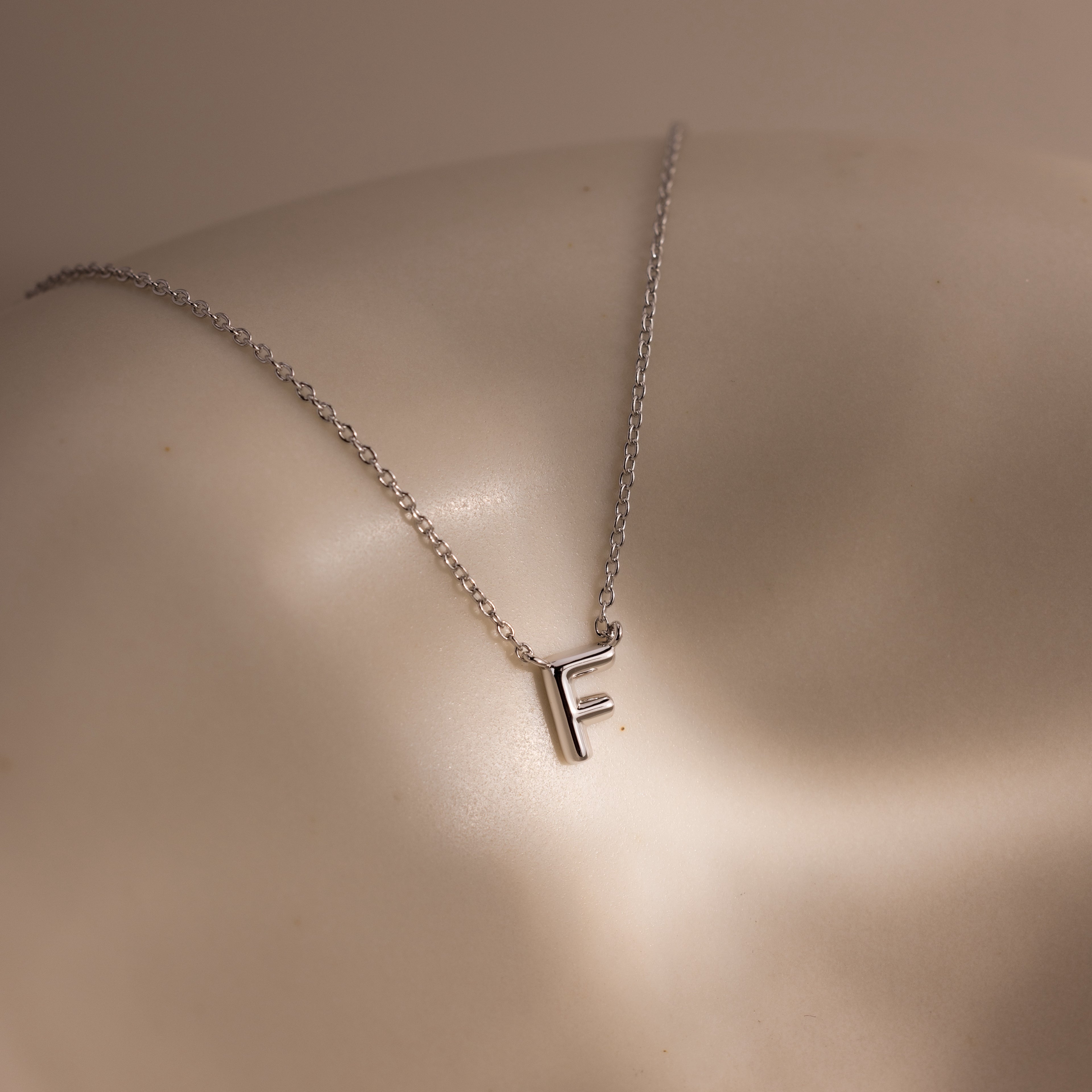 The Dainty Bubble Letter Necklace in Sterling Silver, featuring an F pendant, rests on smooth, softly lit skin—adding a touch of personalized elegance.