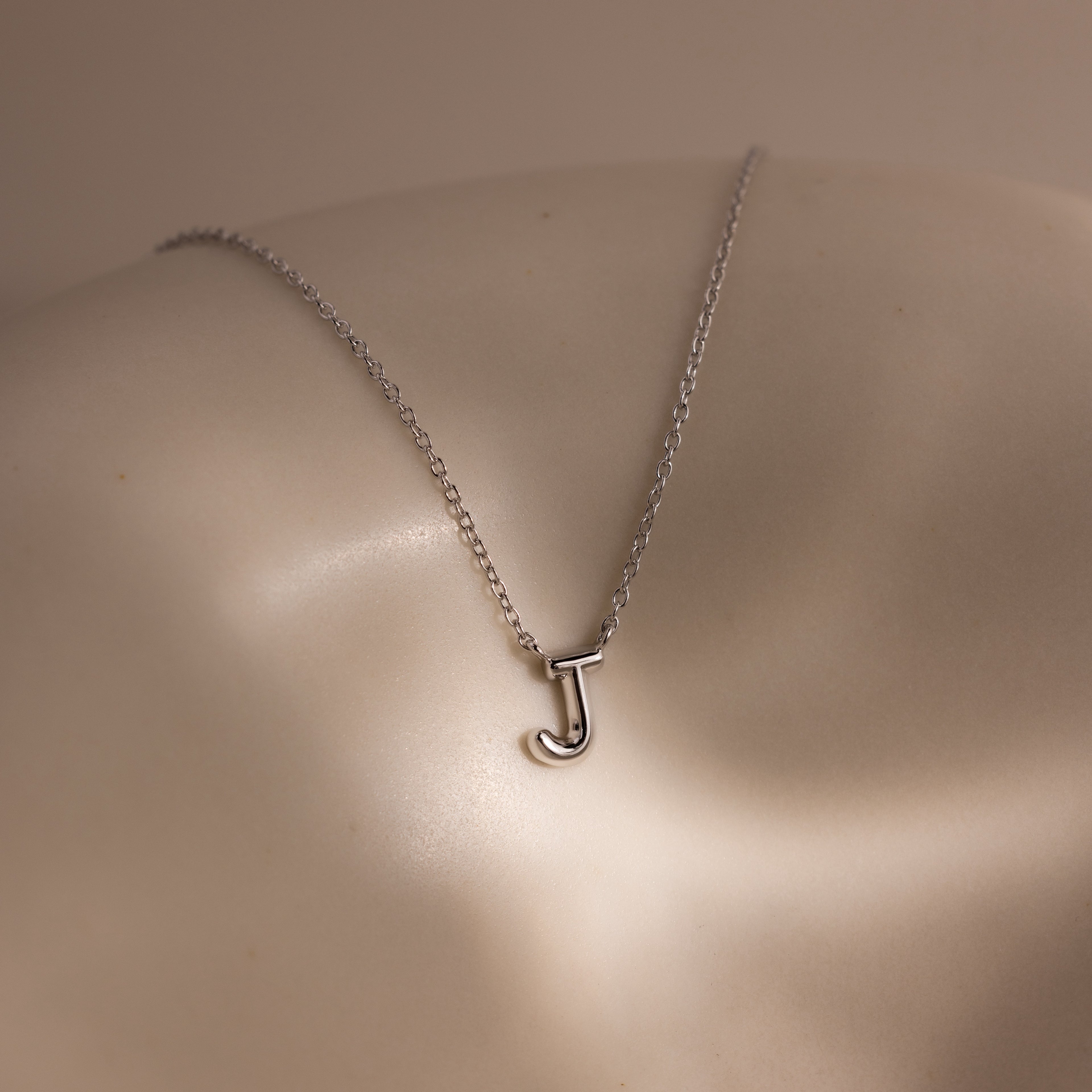 The Dainty Bubble Letter Necklace in Sterling Silver, featuring a J pendant, shines softly against light skin—an ideal personalized accessory for a minimalist style.