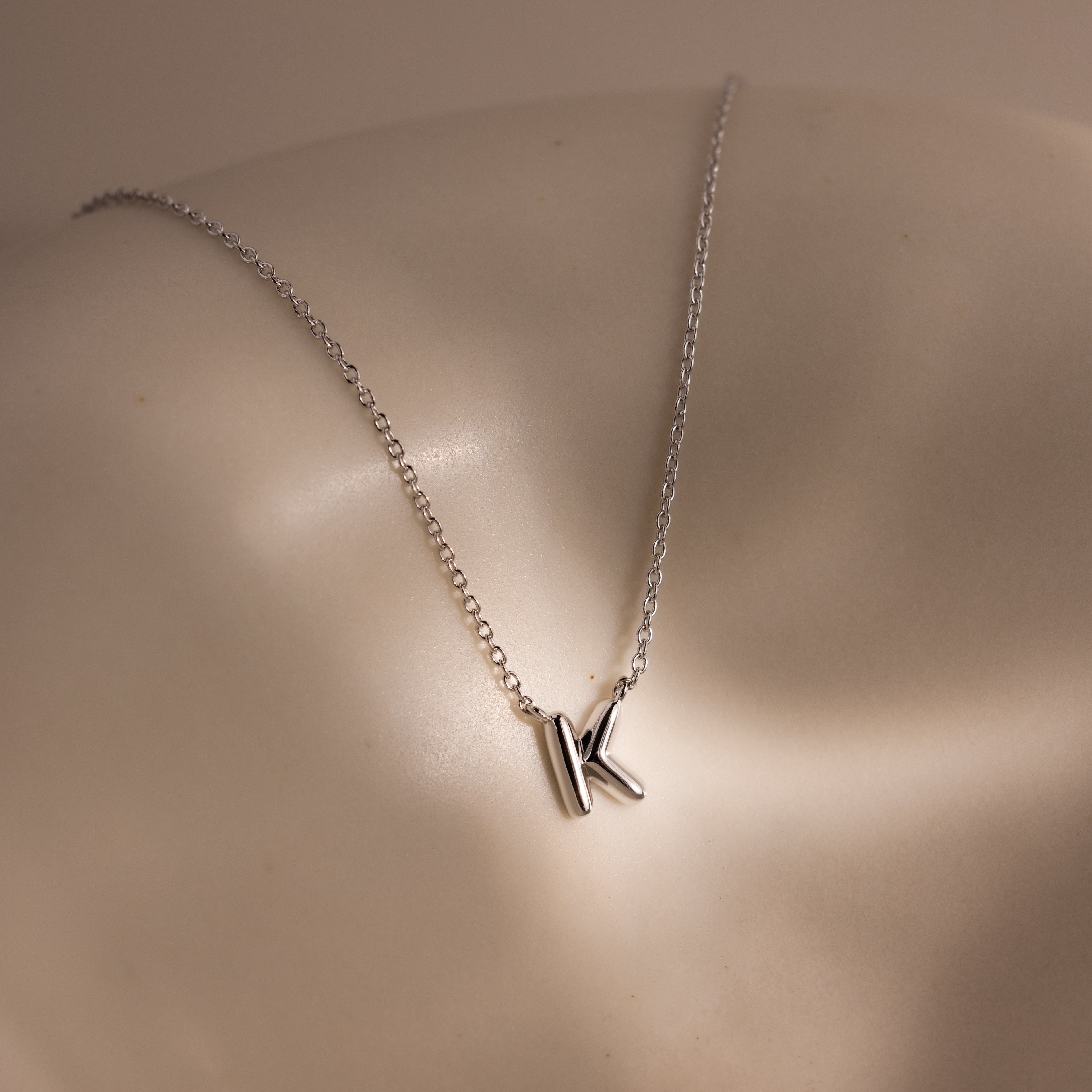 The Dainty Bubble Letter Necklace in Sterling Silver features a K-shaped pendant, resting on a smooth, light-colored surface to add personalized style to any minimalist look.