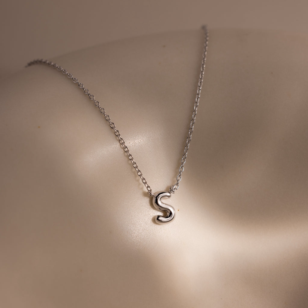 The Dainty Bubble Letter Necklace in Sterling Silver features a minimalist silver S pendant, resting elegantly on pale skin under soft lighting.
