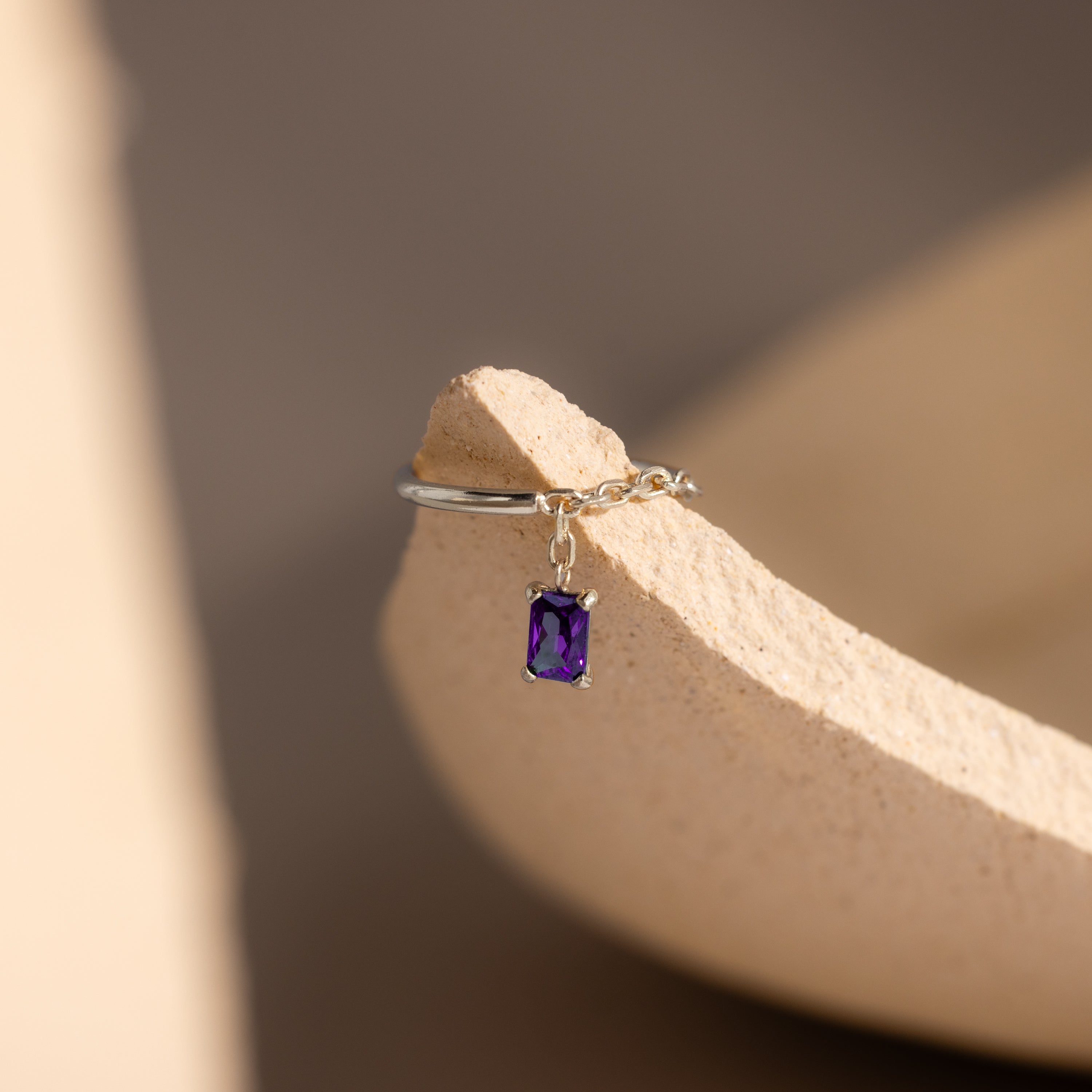 The Amethyst Drop Chain Ring, featuring a silver chain band and a dangling emerald-cut purple gemstone, rests on a textured beige surface.